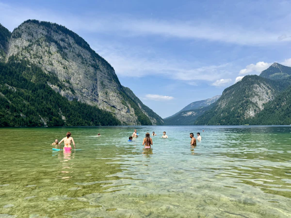 Teen travelers wade and swim in clear green water below steep mountain walls at the Königssee