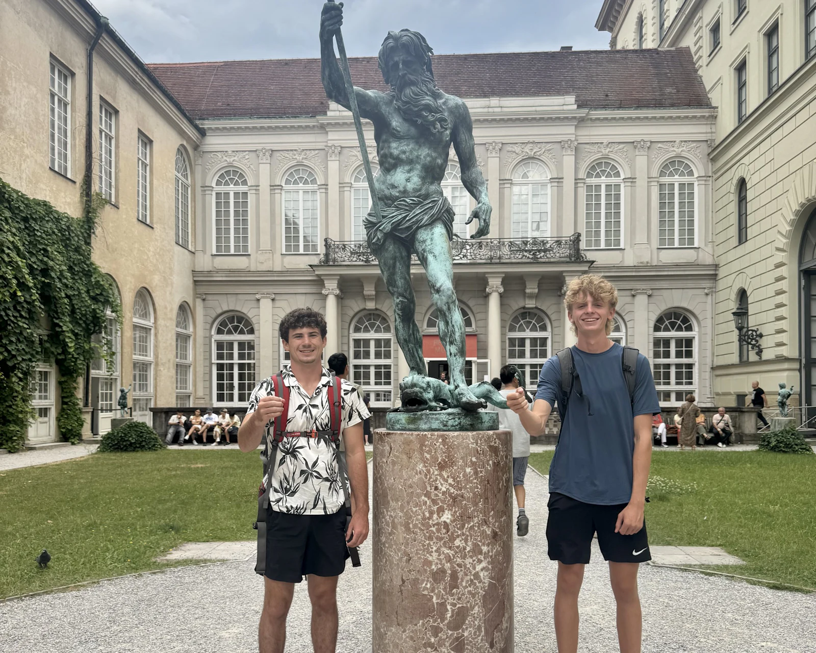 Two teen travelers stand on either side of a bronze statue fountain in a palace courtyard in Munich