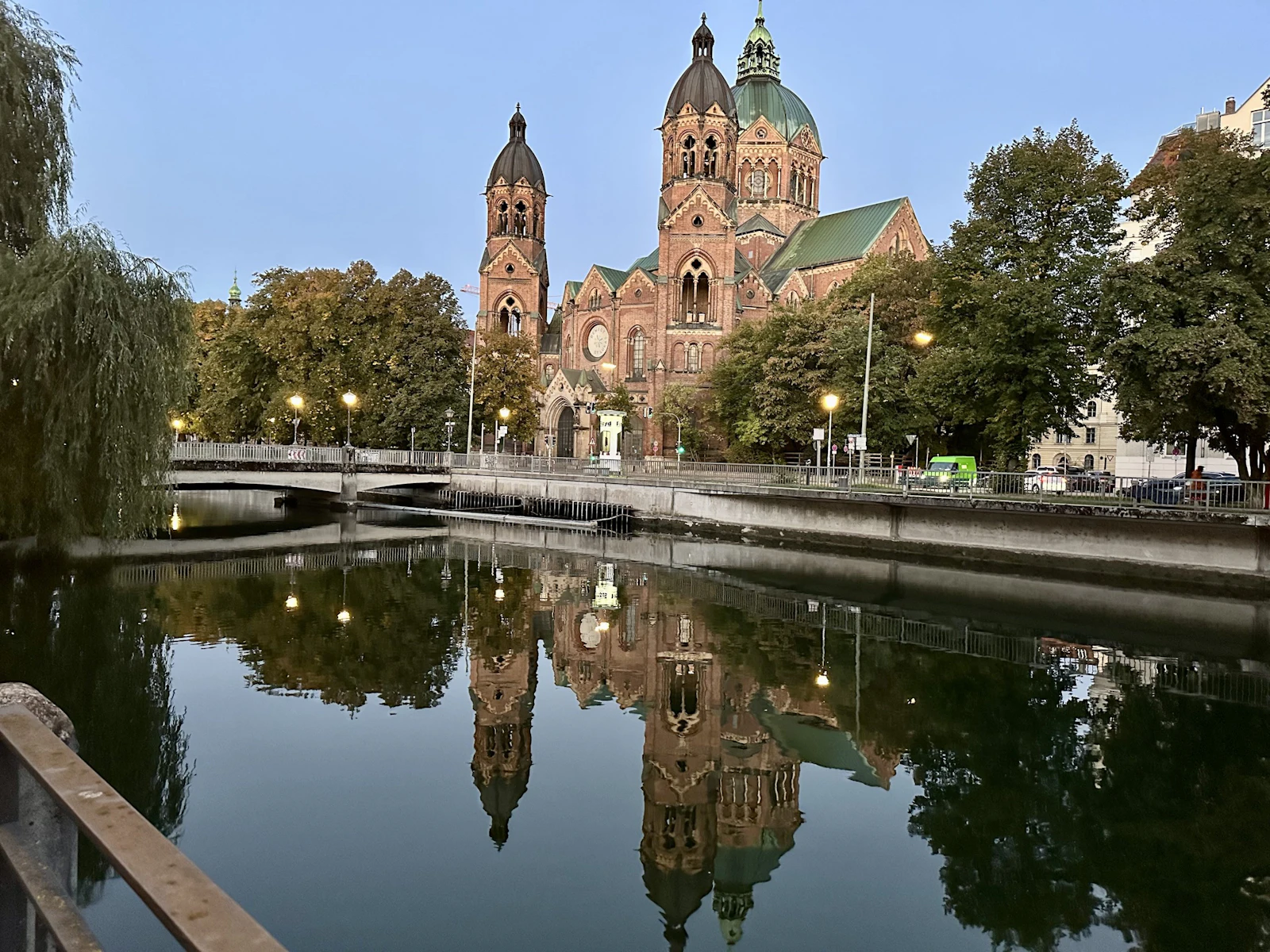 A red-brick church with green domes is reflected in the still water of the Isar in Munich