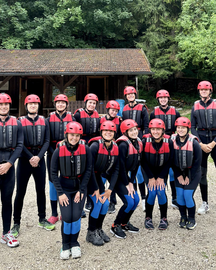 Teen travelers in wetsuits, red helmets and life vests line up beside a wooden hut before rafting
