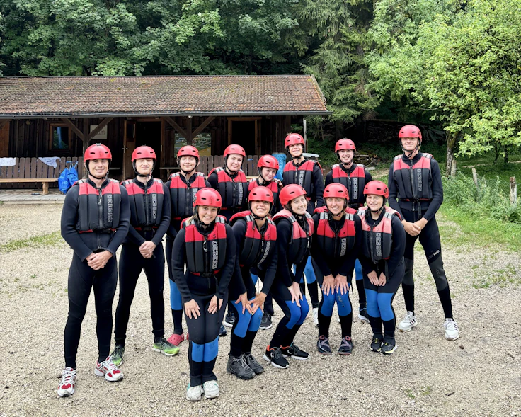 Teen travelers in wetsuits, red helmets and life vests line up beside a wooden hut before rafting