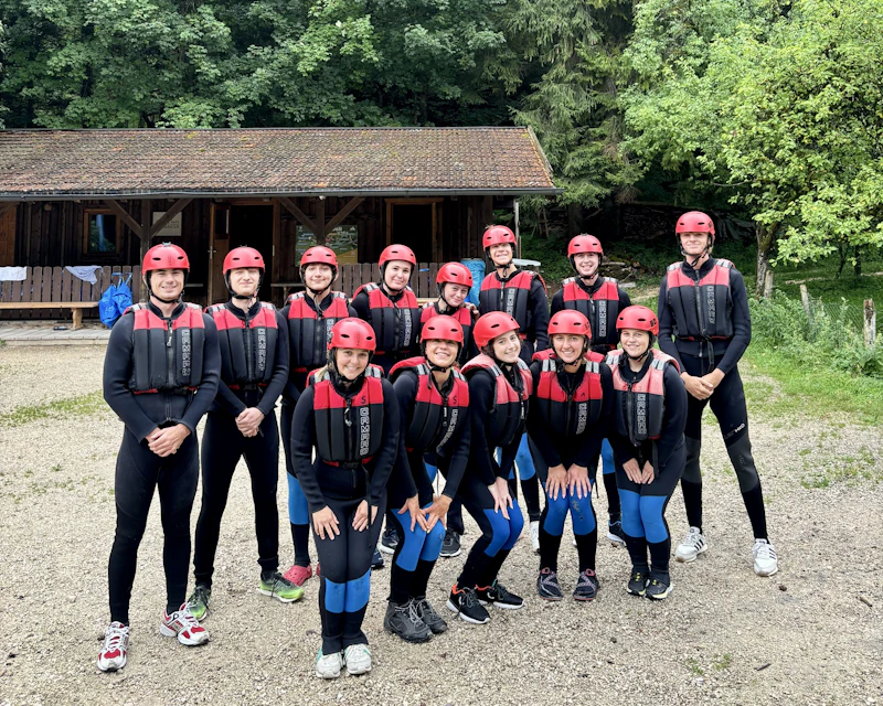 Teen travelers in wetsuits, red helmets and life vests line up beside a wooden hut before rafting