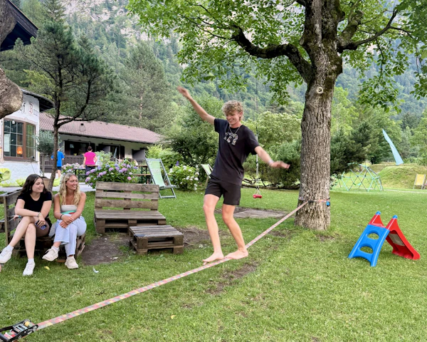 A teen traveler balances on a slackline strung between trees on a lawn while friends watch, mountains behind