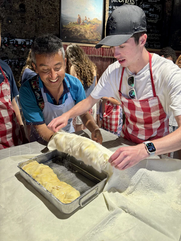 A teen traveler and an adult in aprons roll apple strudel dough into a baking pan during a cooking class