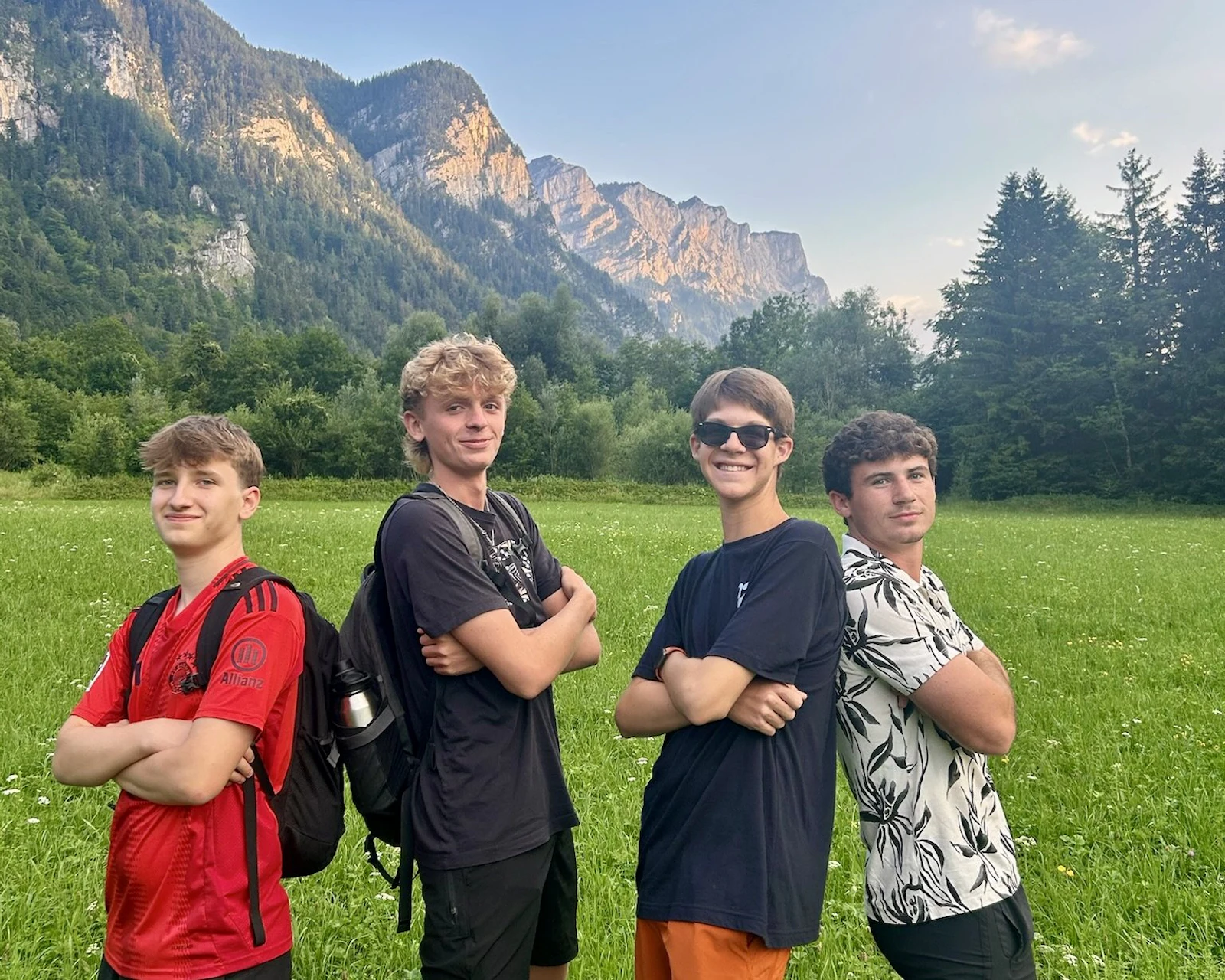 Four teen travelers pose with folded arms in a green meadow below limestone peaks