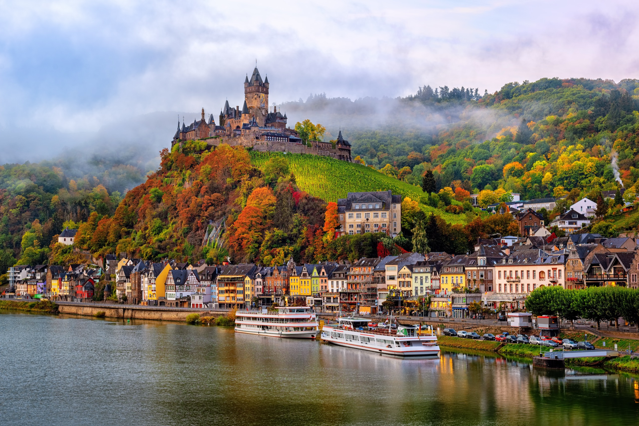 A turreted hilltop castle above a riverside German town in autumn, with pastel-colored houses along the waterfront, two white river cruise boats moored below, forested hillsides in autumn color, and low morning mist.