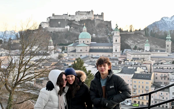 Three students in winter coats and hats stand at a viewpoint above Salzburg, with the hilltop fortress, the cathedral domes and snow-dusted mountains behind them.