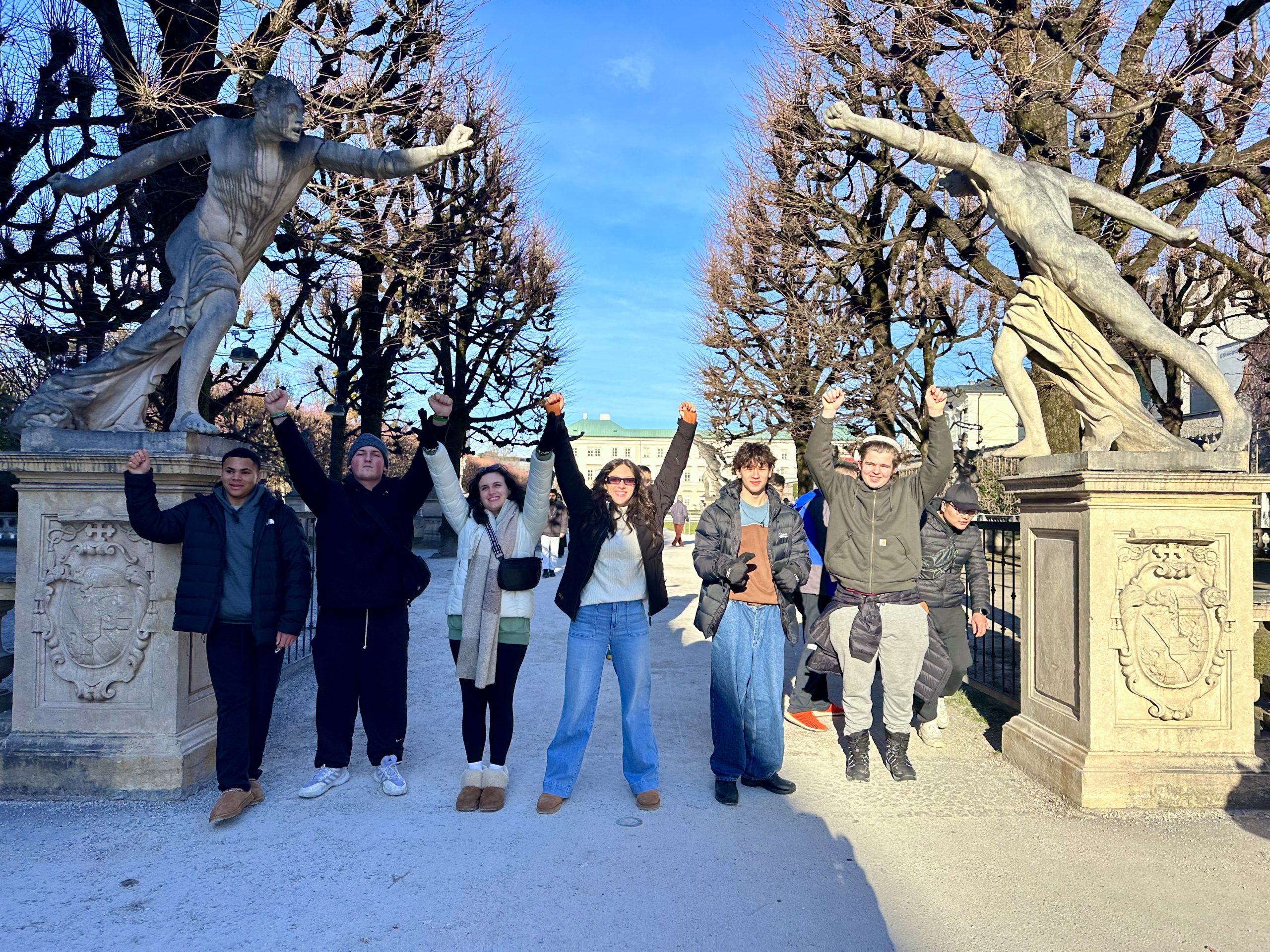 Group of teen travelers celebrating in Mirabell Gardens, Salzburg, Austria, with statues and winter trees behind them on a student travel adventure.