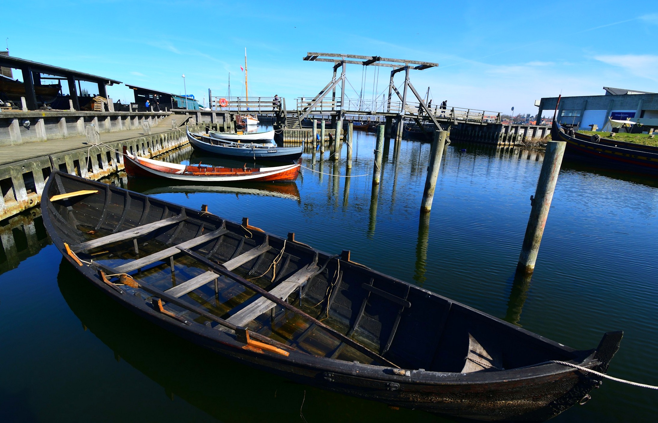 A clinker-built wooden rowing boat sits low in the still water of Roskilde harbor, a small lifting bridge behind it.