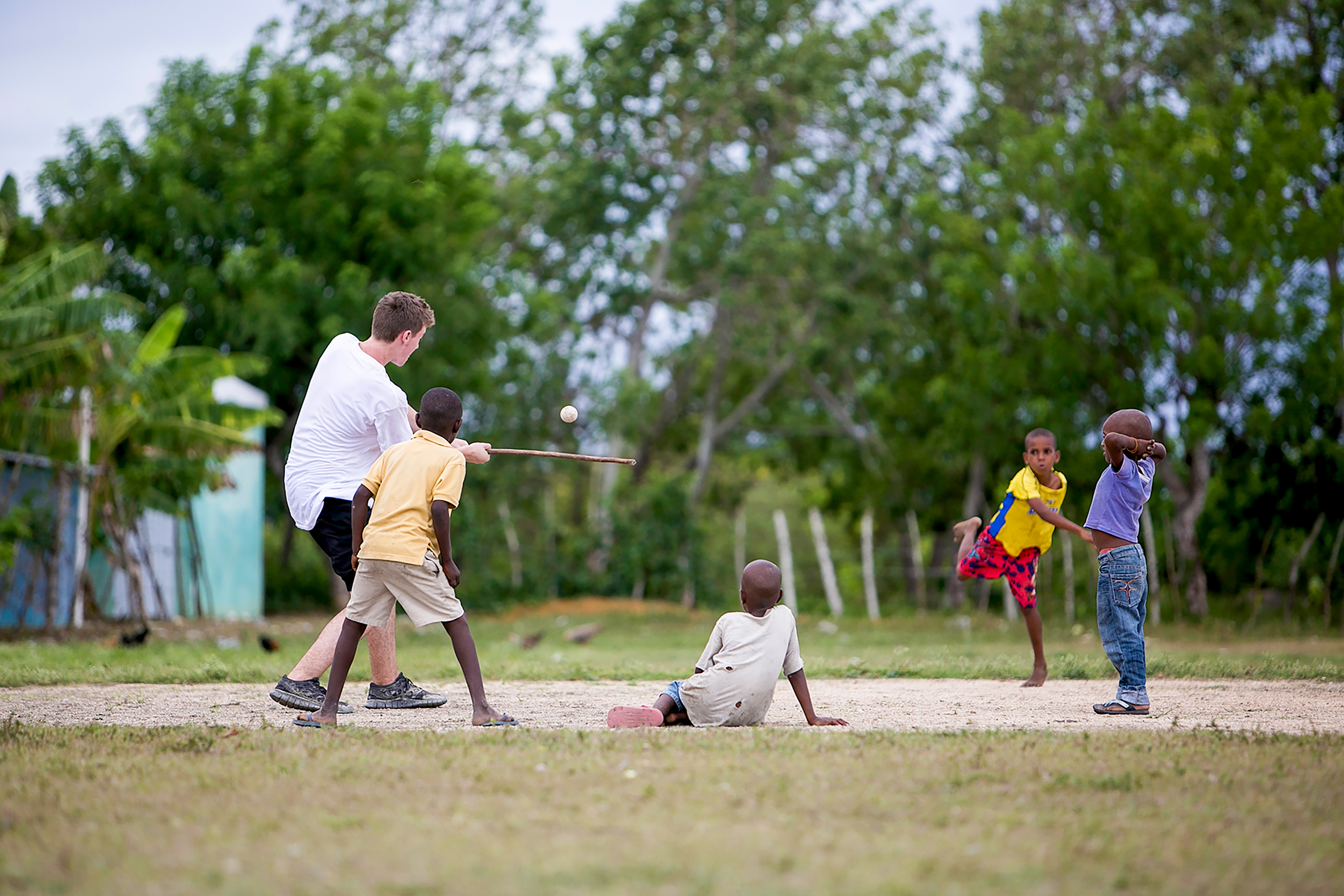 A student pitches a ball while local children play baseball on a dirt field