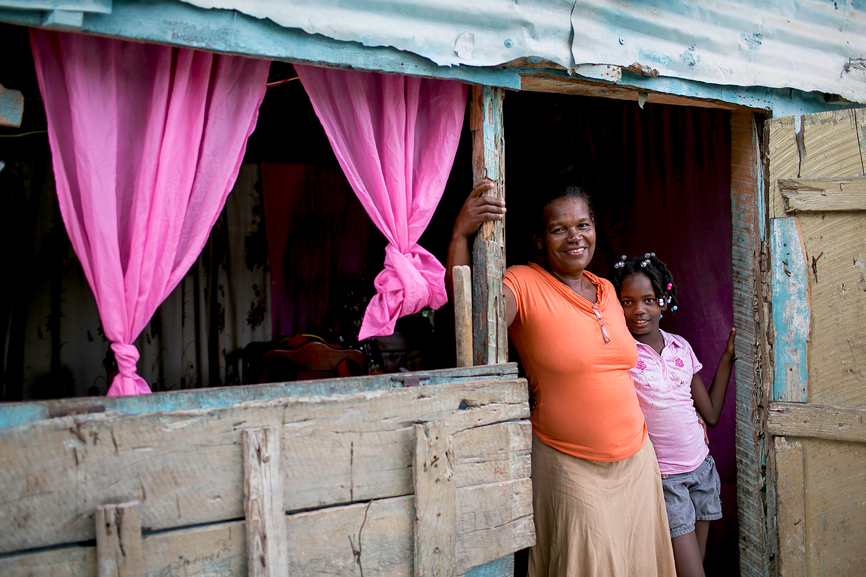 A woman and girl smile in the doorway of a home with pink curtains