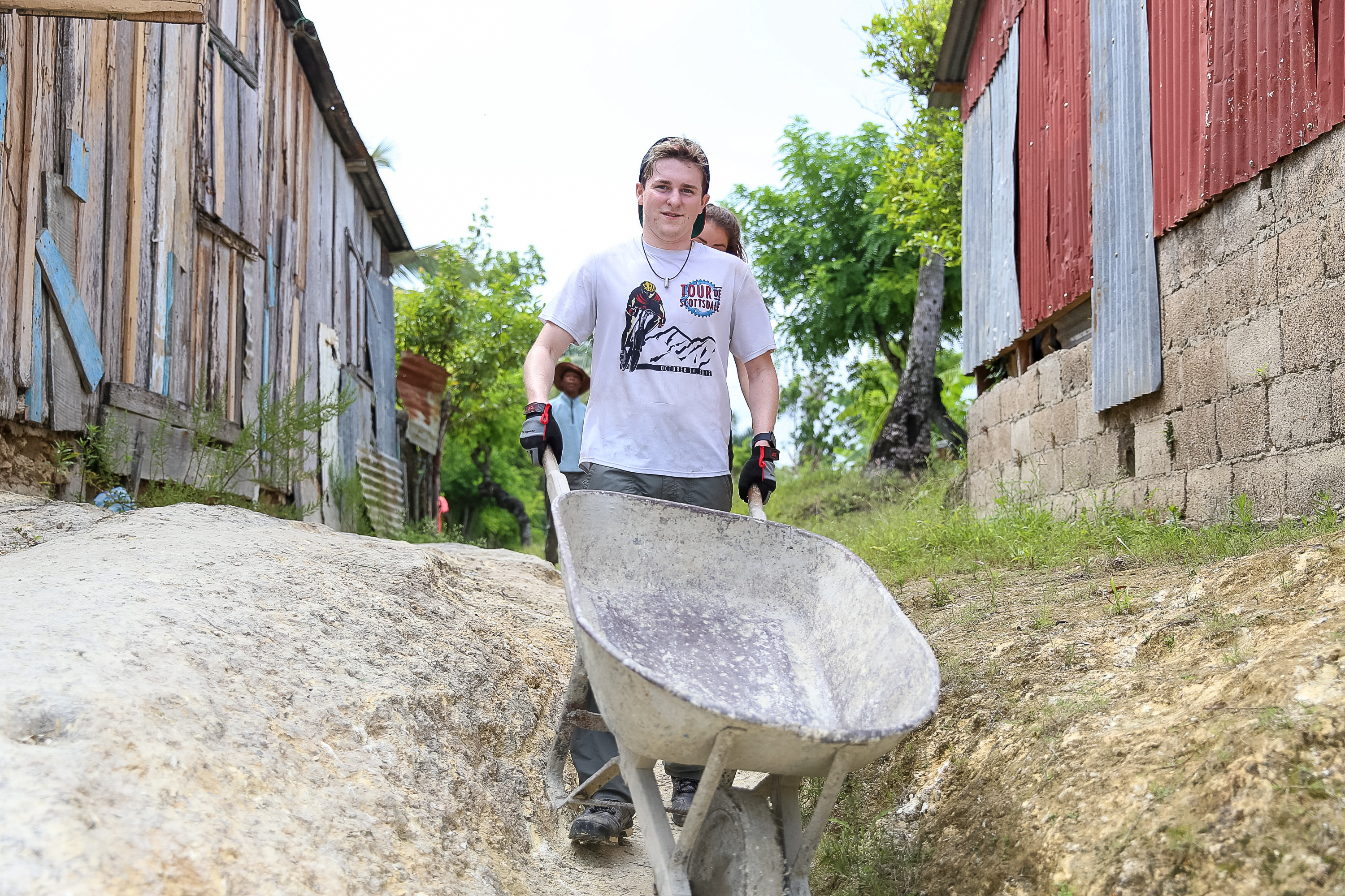 A student pushes a wheelbarrow along a dirt path between wood and tin-roofed houses