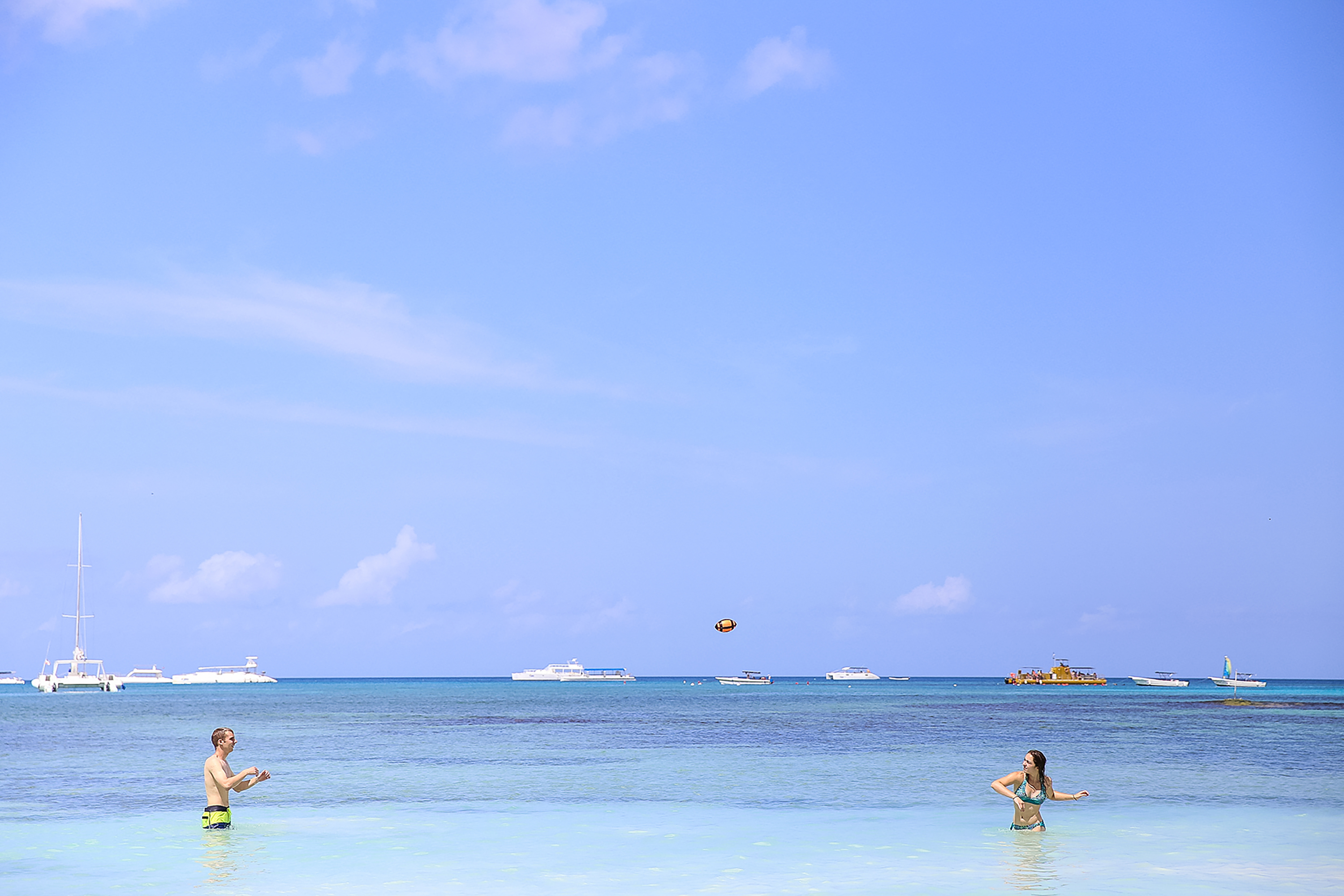 Students wade in shallow turquoise water tossing a ball, boats anchored offshore