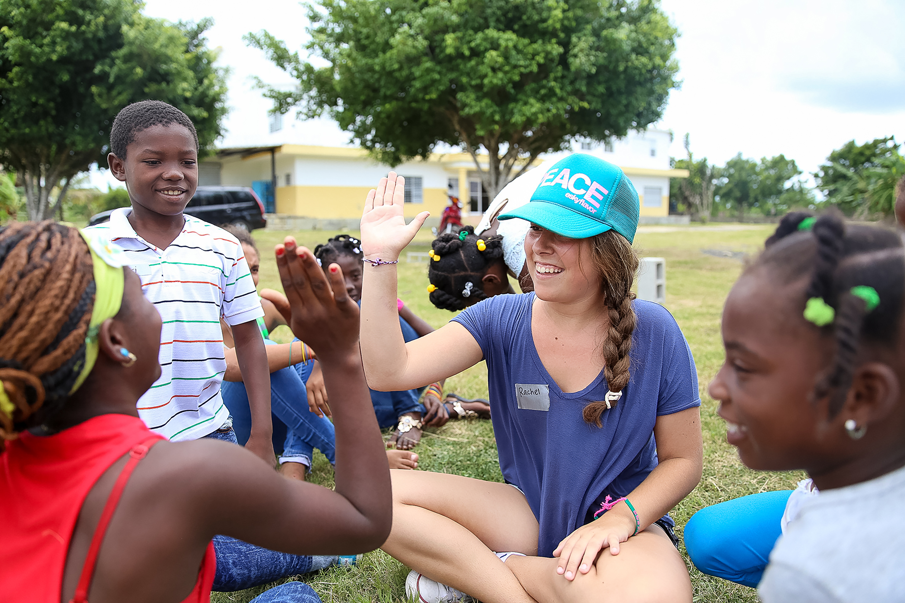 A student high-fives local children while sitting in the grass outdoors
