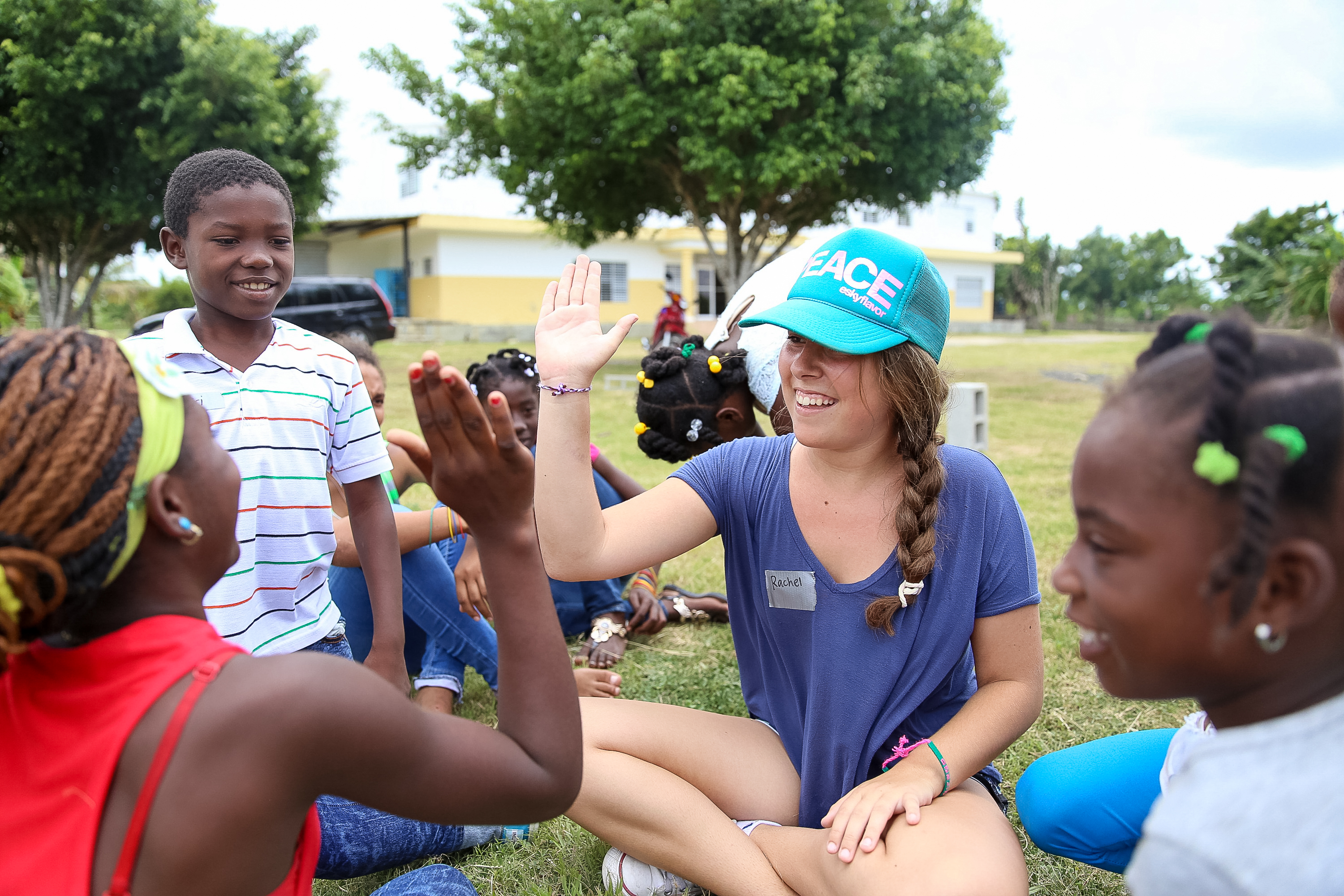 A student in a turquoise cap sits cross-legged on the grass in a circle of local children, trading high fives, with a yellow-and-white building and trees behind them.