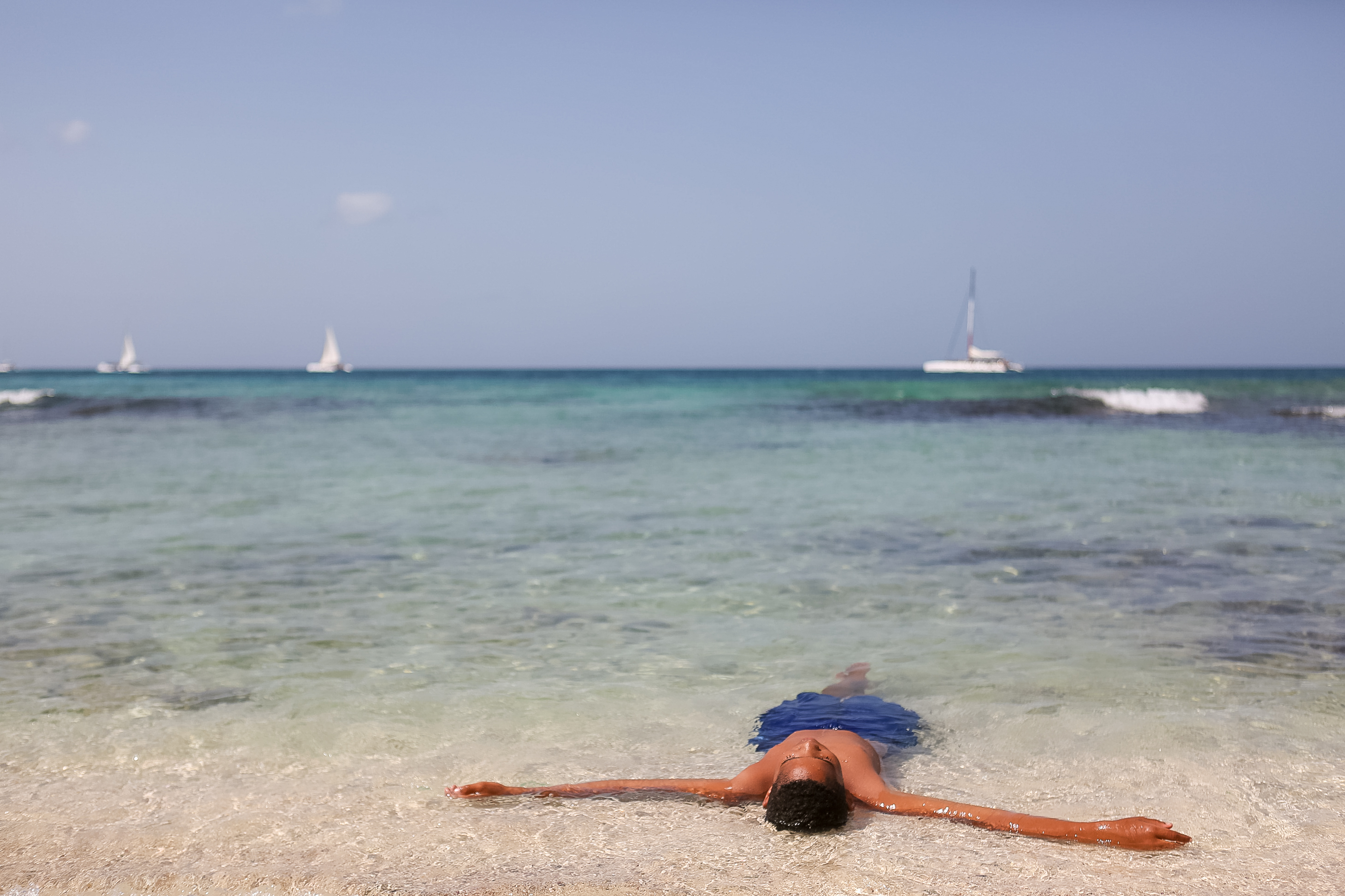 A student floats on their back in shallow turquoise water with sailboats on the horizon