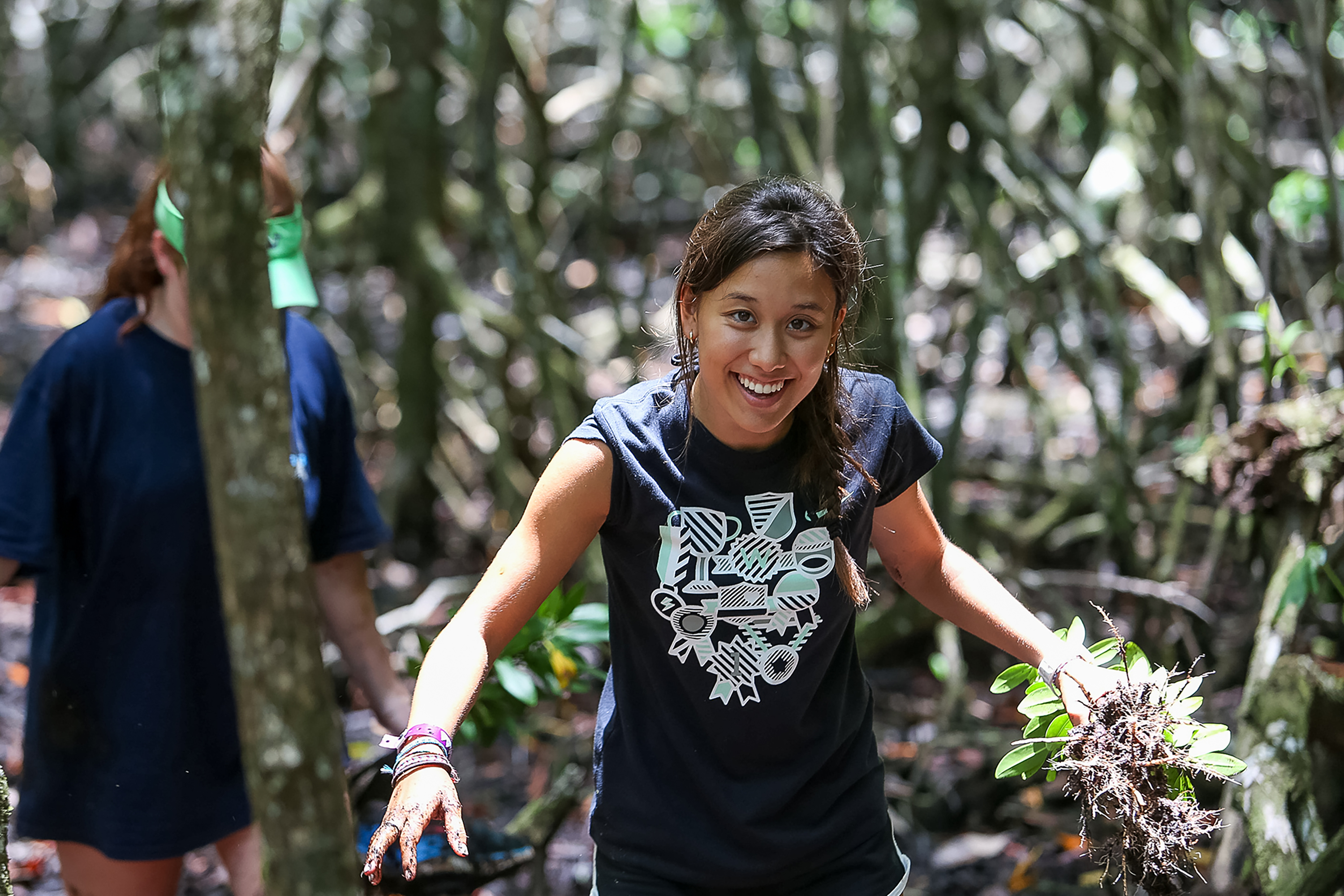 Student carrying plants during mangrove restoration project with trees in the background.