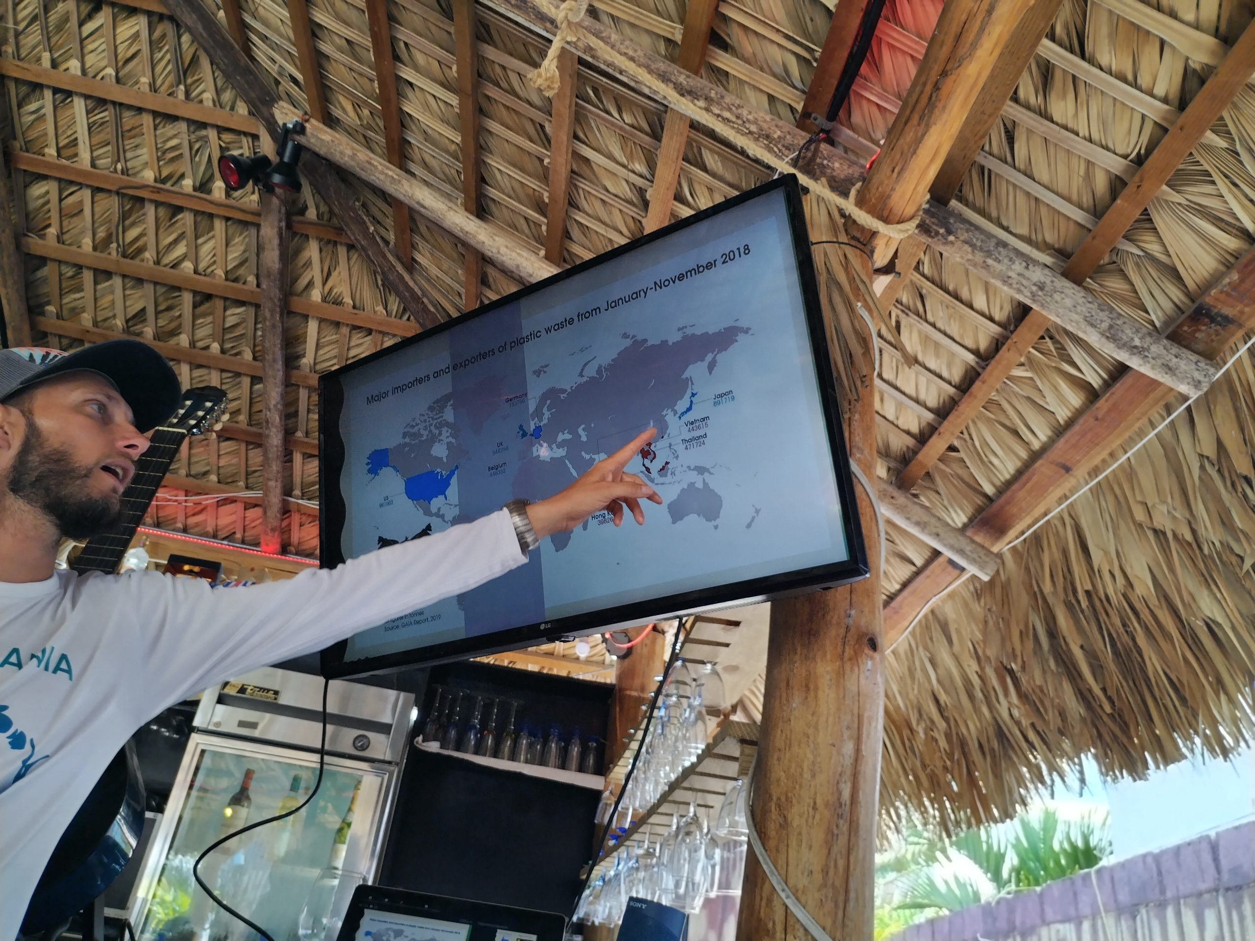 A leader points to a world map on a screen under a thatched roof