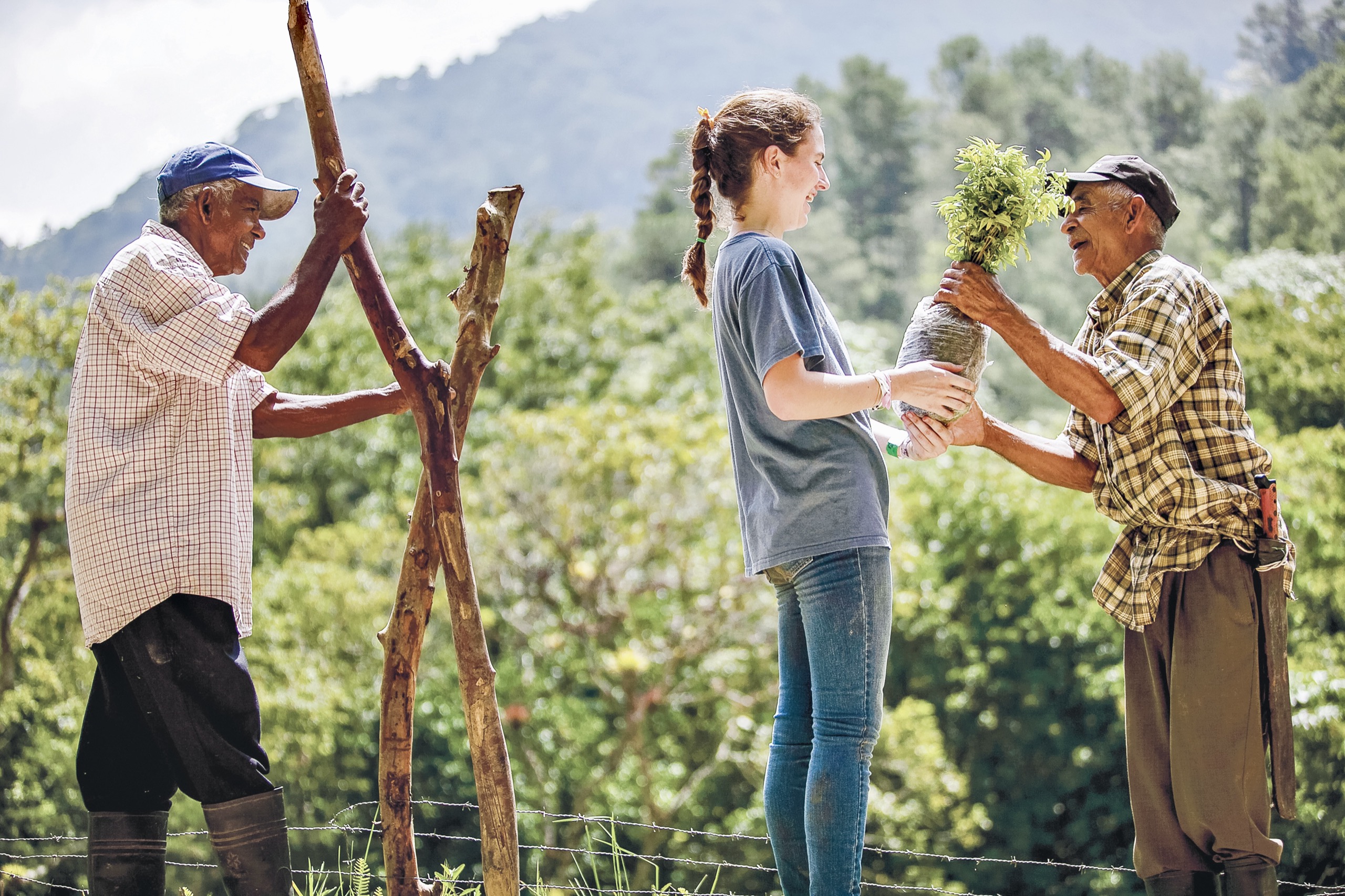 A student in a grey t-shirt and jeans hands a wrapped seedling to an older man in a plaid shirt and flat cap, while a second older man in a blue cap and checked shirt braces two crossed wooden stakes, with a barbed-wire fence, green hillside and a mountain ridge behind them.