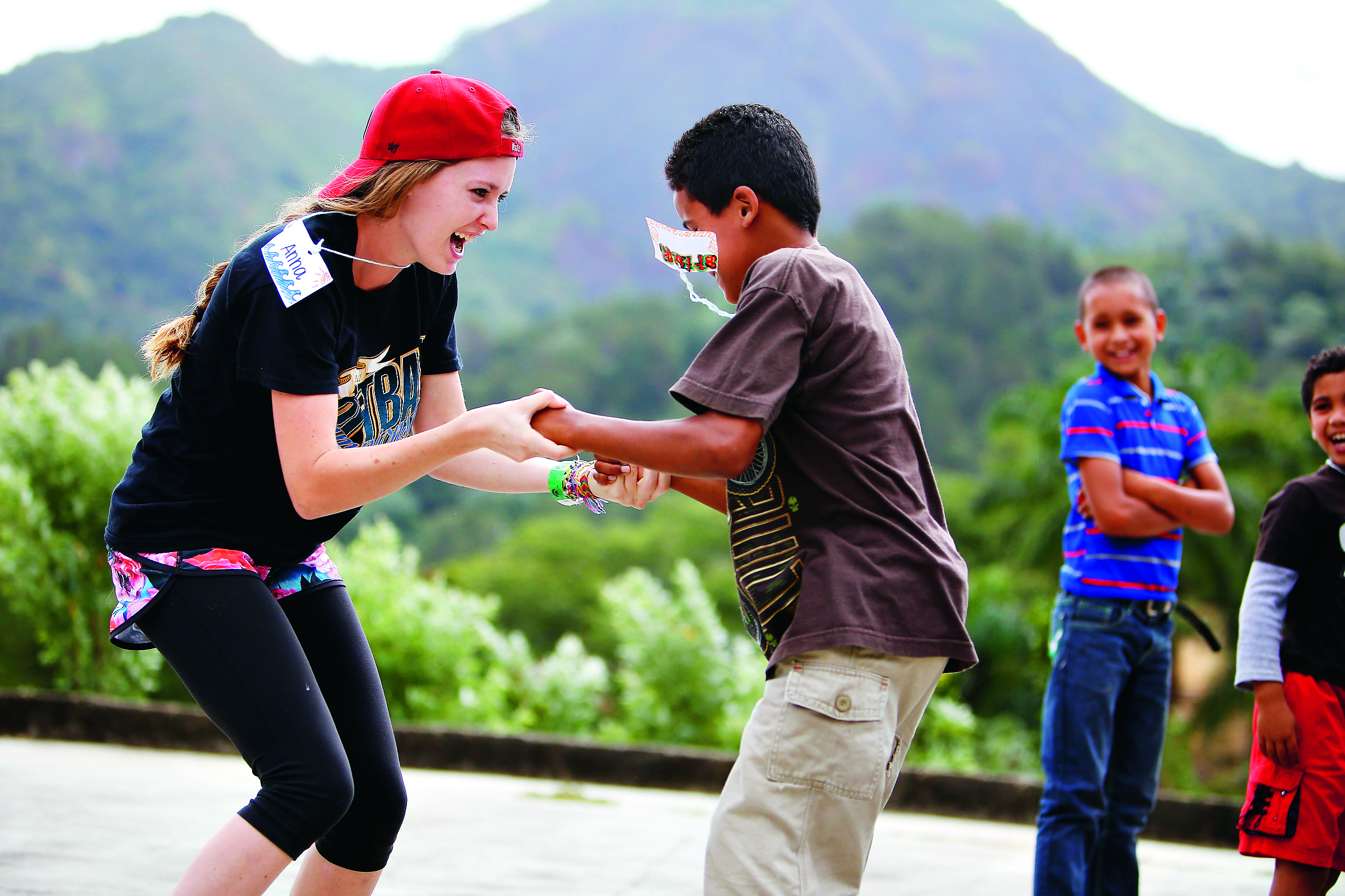 A student in a red cap laughs while clasping hands with a local boy wearing a paper mask, as two other boys watch and smile, with a green mountain ridge behind them.