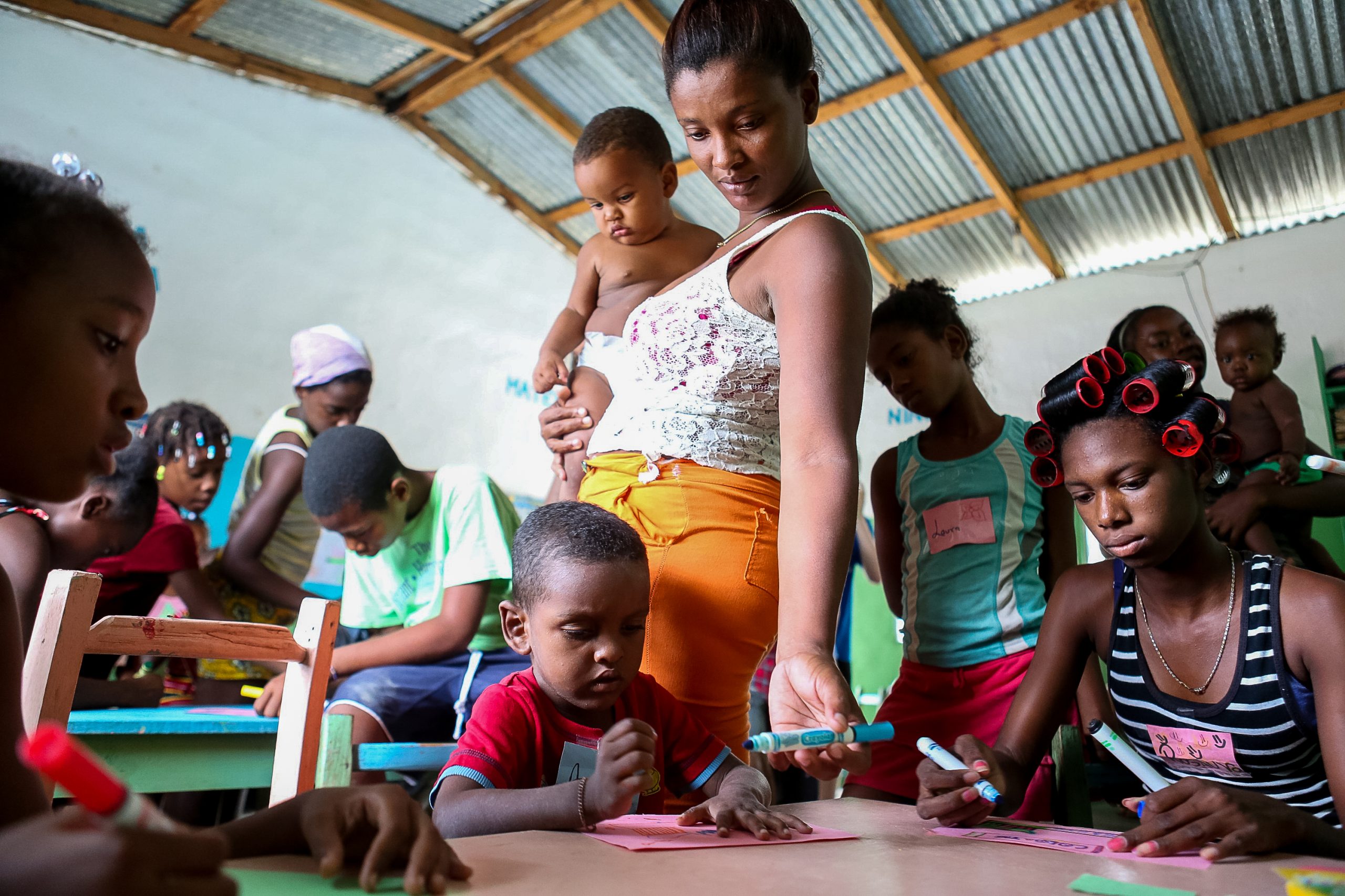 Community members color at a table under a tin-roofed shelter while a woman holds a baby