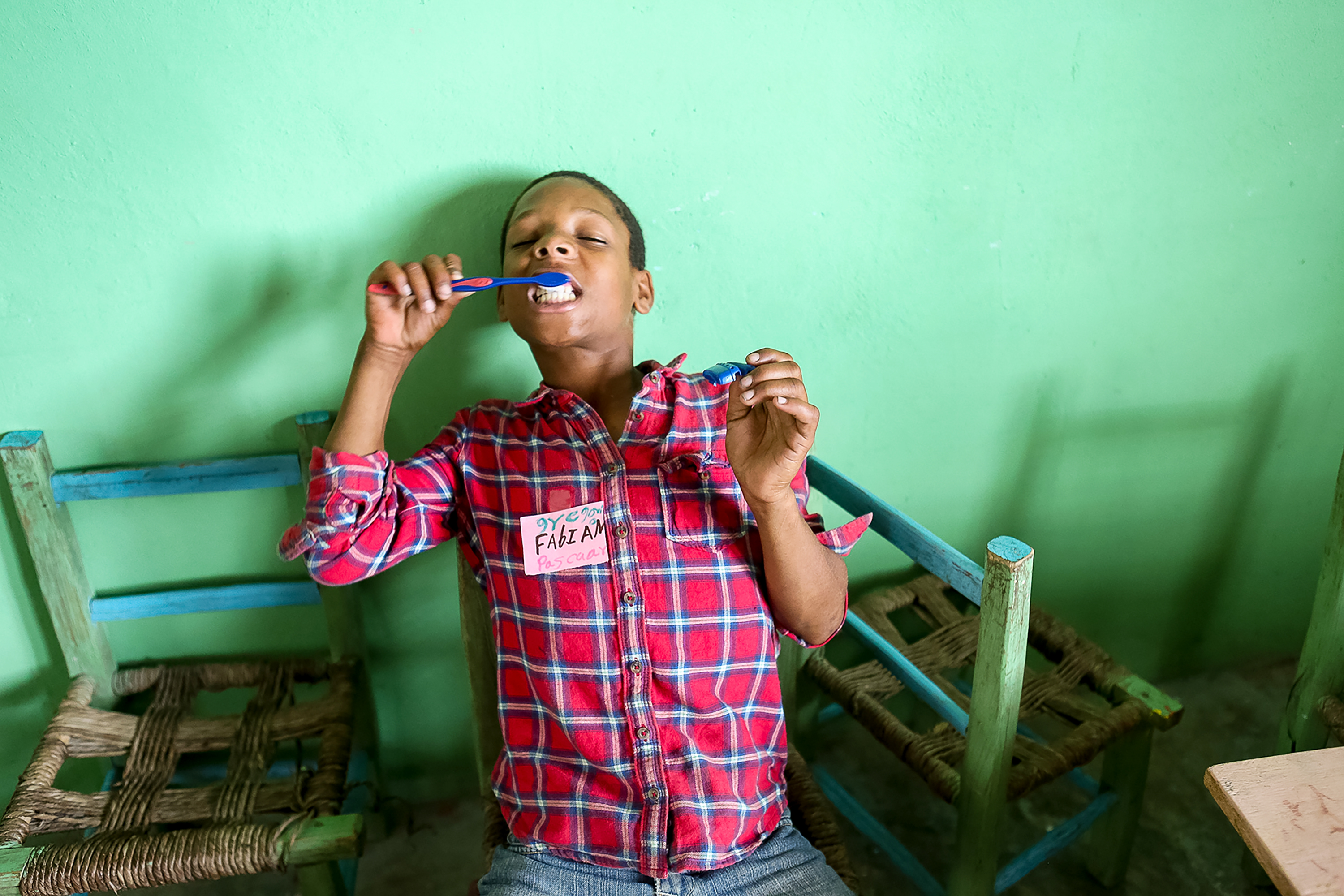 A young child brushes their teeth while seated between wooden chairs