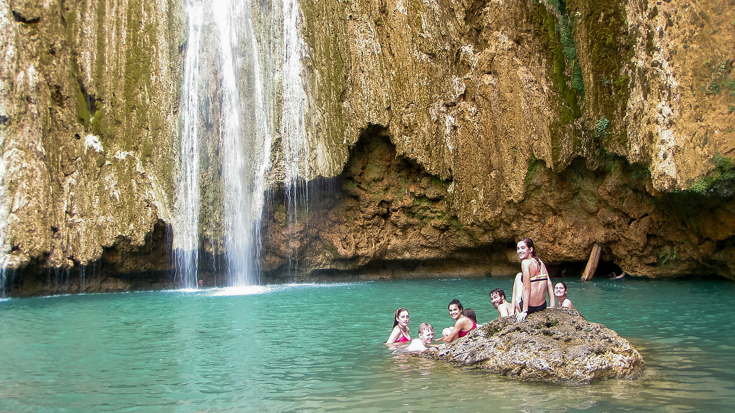 Students gather on a rock in a turquoise pool beneath a waterfall