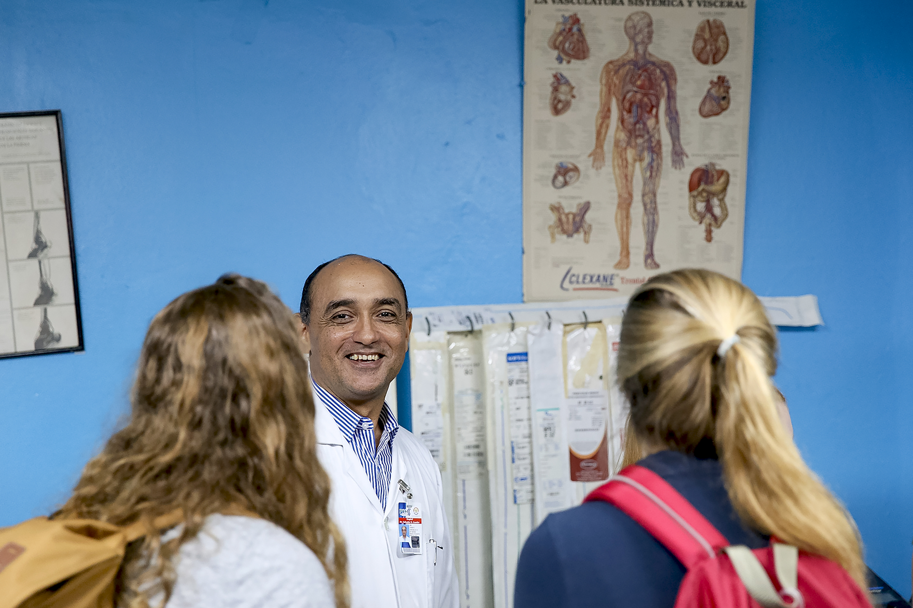 A doctor smiles while talking with students in a clinic room with an anatomy poster on the wall