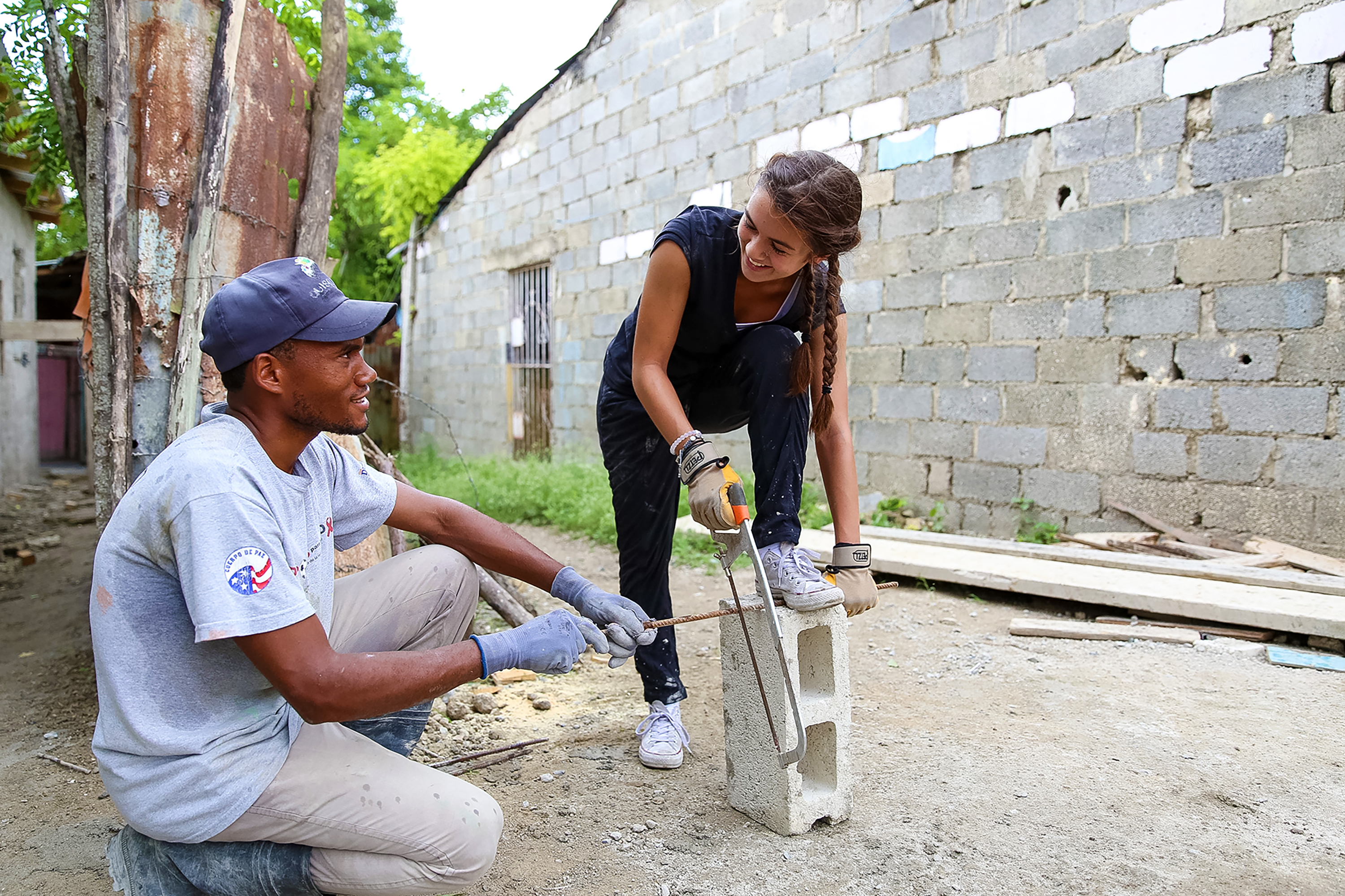 A student uses a saw on a cinder block while a local worker crouches nearby on a construction site