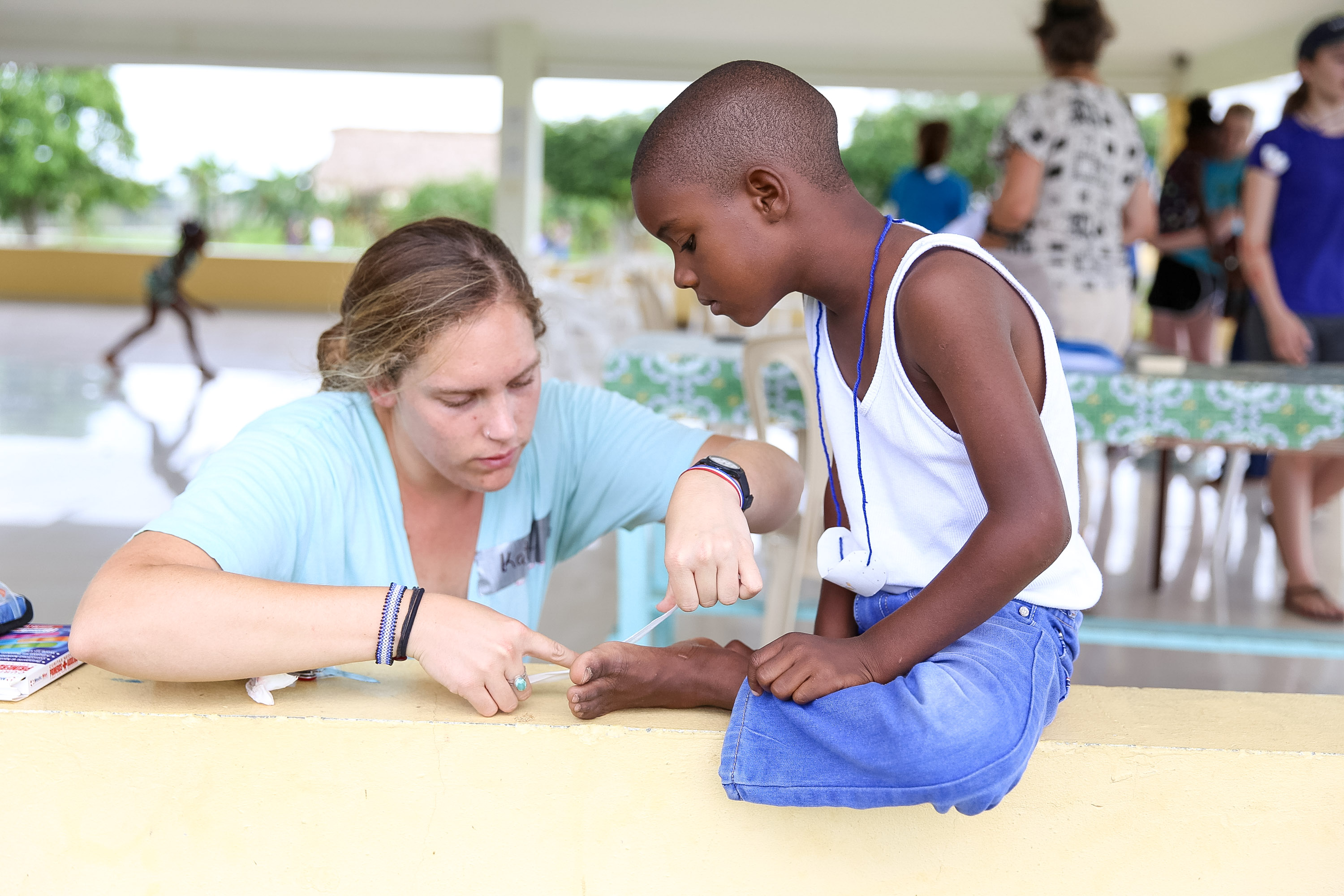A student sits at a wall painting a young boy's fingernail during a community health activity