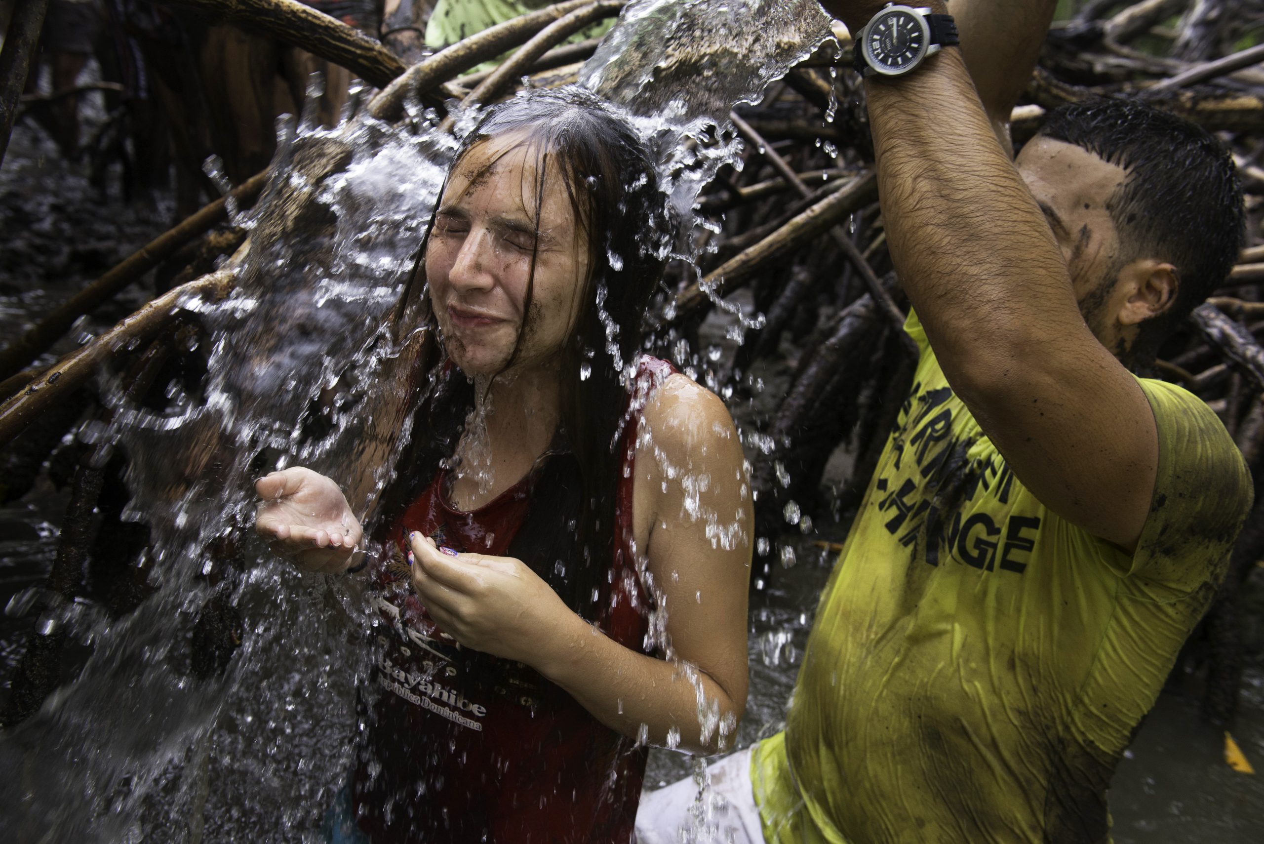 a girl being drenched in water by a guy behind in a yellow t-shirt. They seem to be out in nature with some brown trees around them.