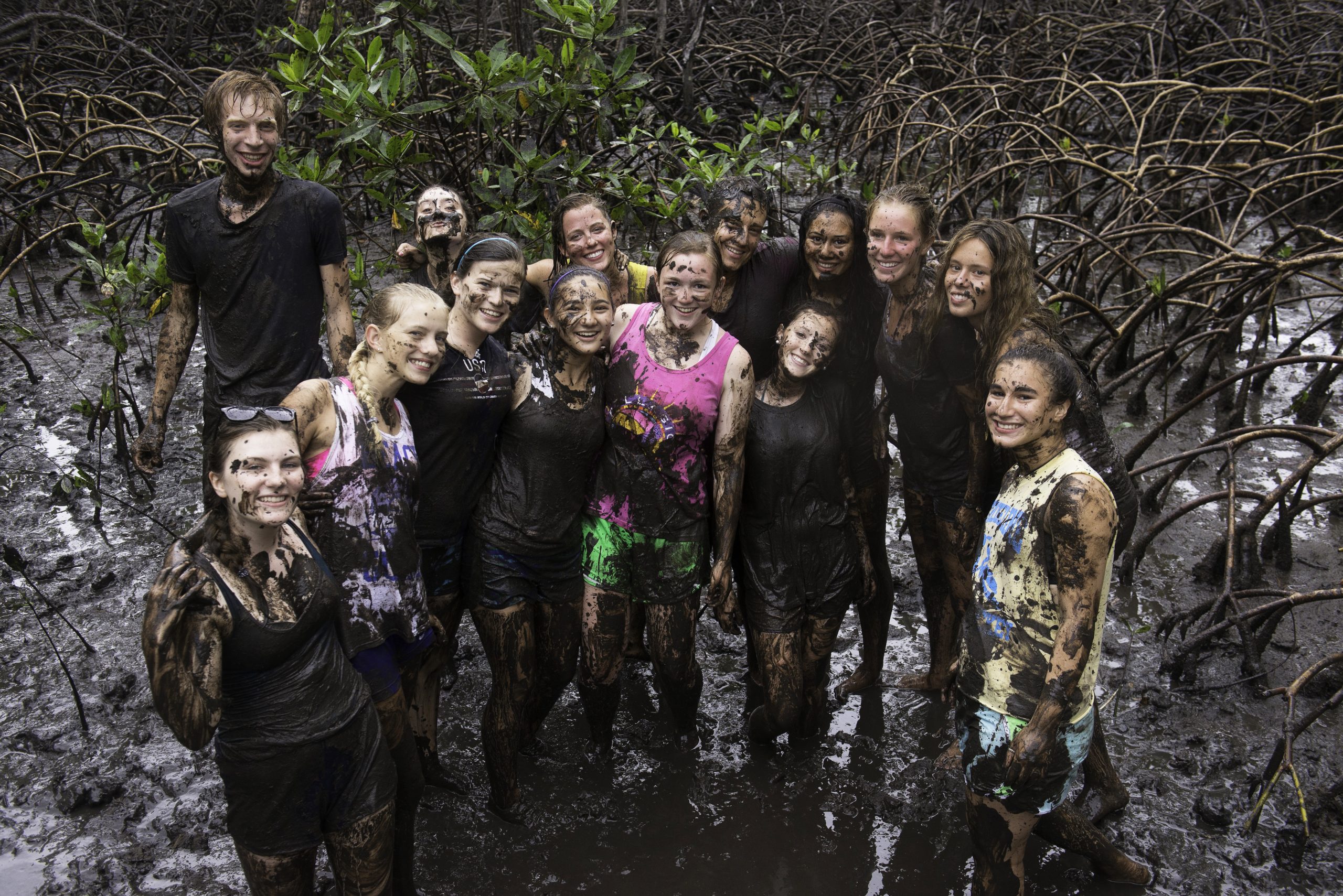 a group of students outside covered in mud but still standing and smiling for the photo that is taken slightly from above.