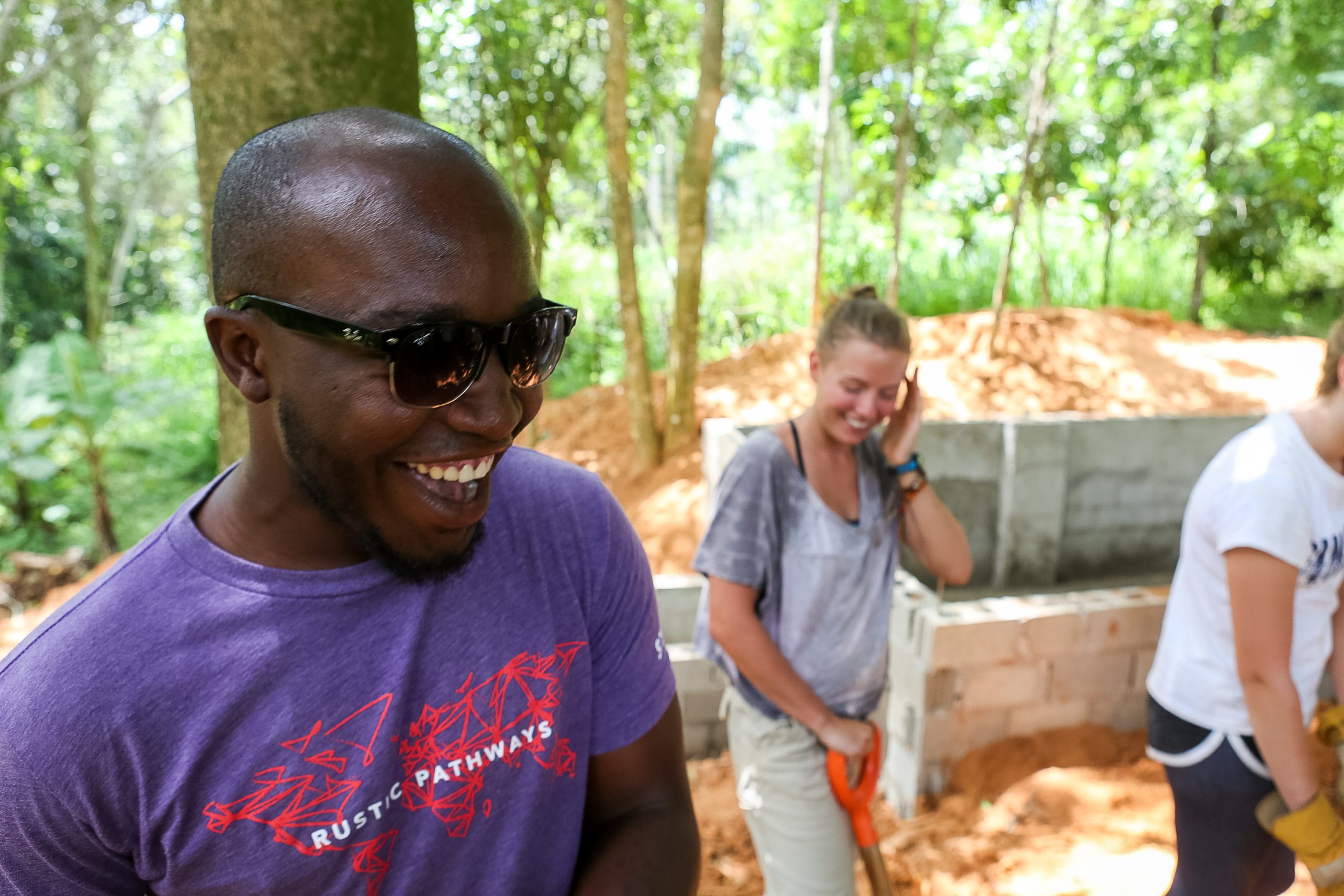 a smiling guy with white teeth and sunglasses wearing a purple t-shirt, face pointing down in the right direction.