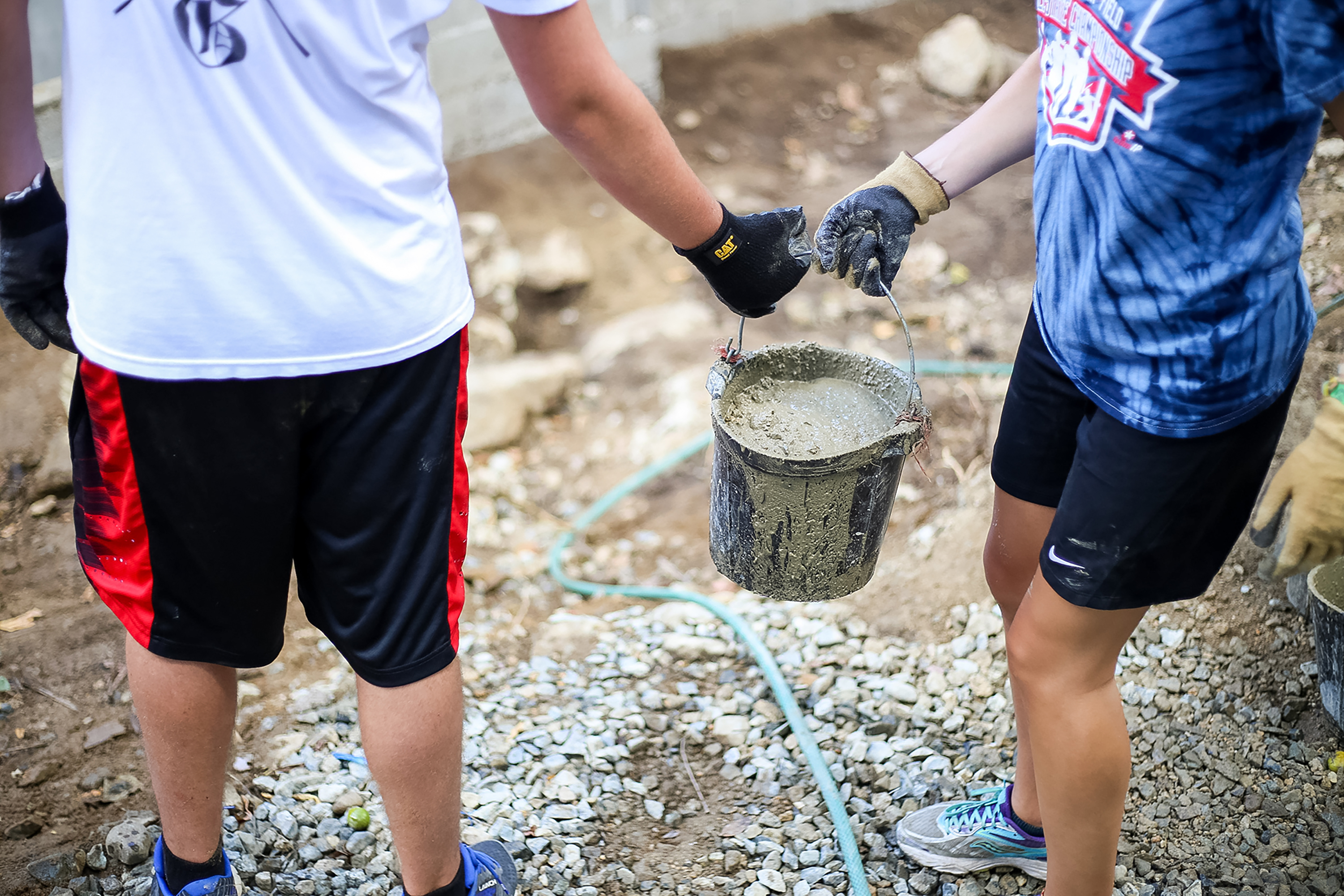 Students wearing work gloves carry a bucket of wet cement on a service site