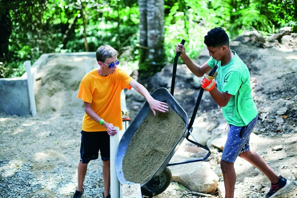 Two teen travelers load sand into a wheelbarrow at a service project construction site in the Dominican Republic.