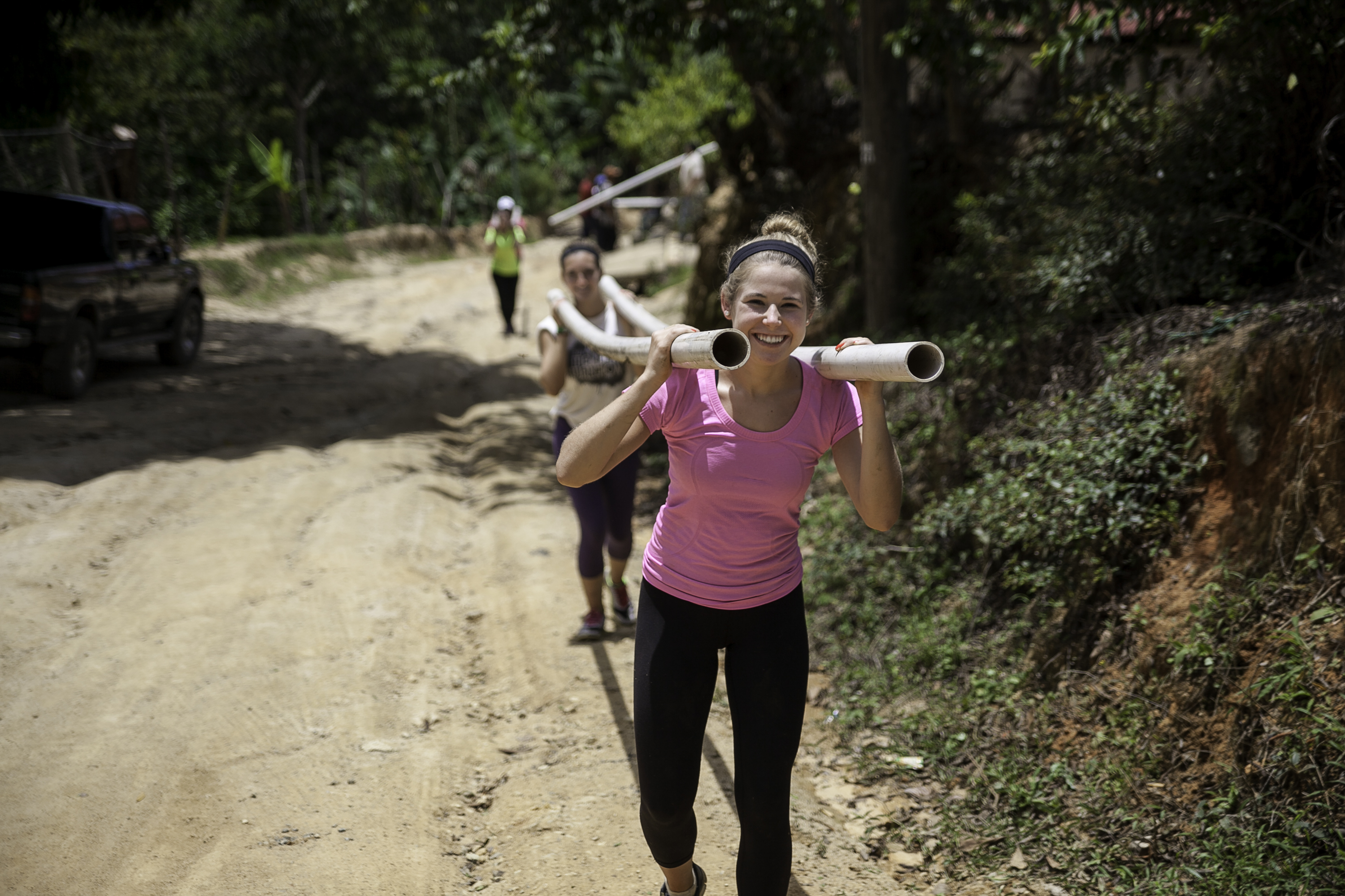 Students carry PVC pipes on their shoulders along a dirt road for a community service project