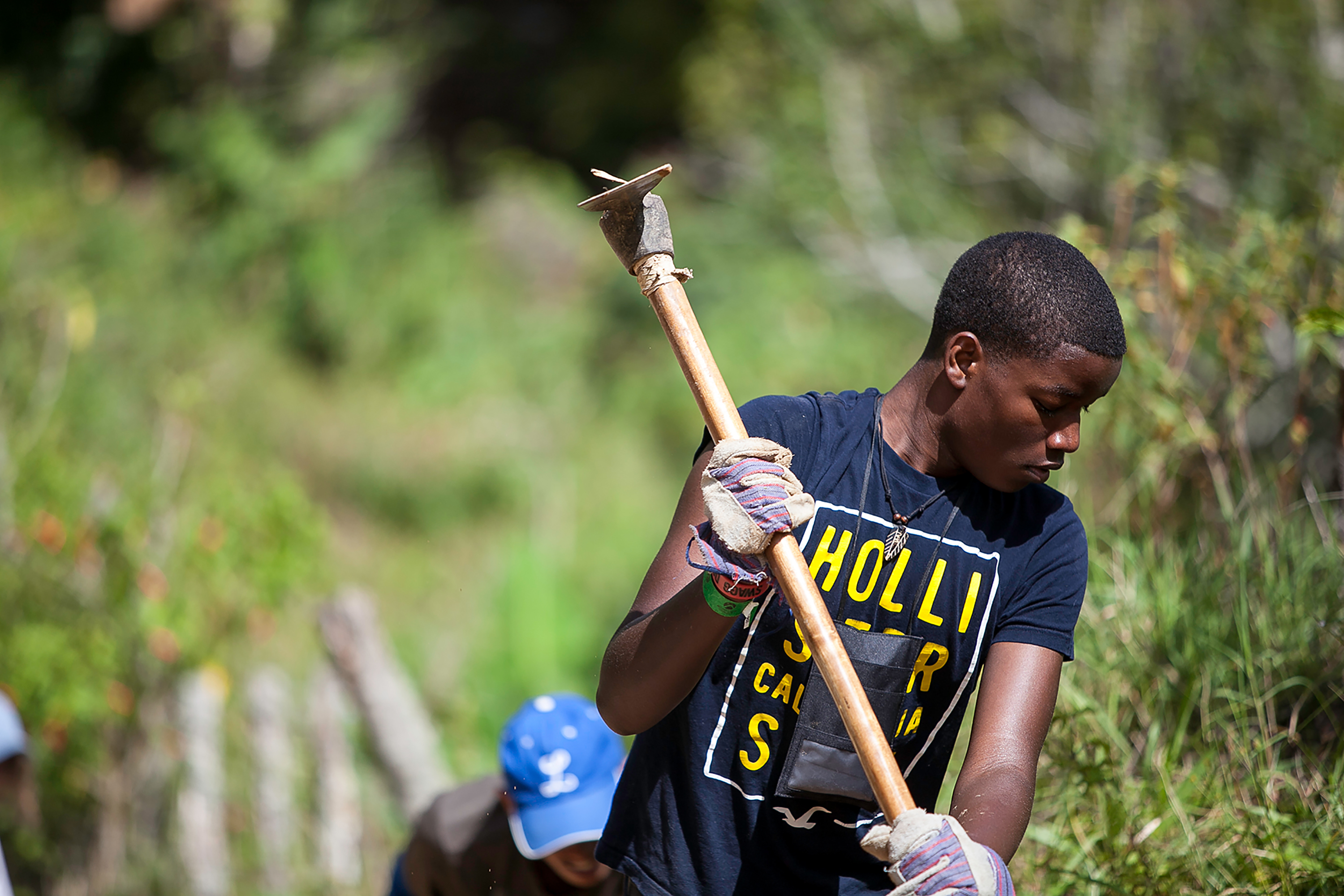 A student wearing work gloves lifts a tool along a trail during a service project