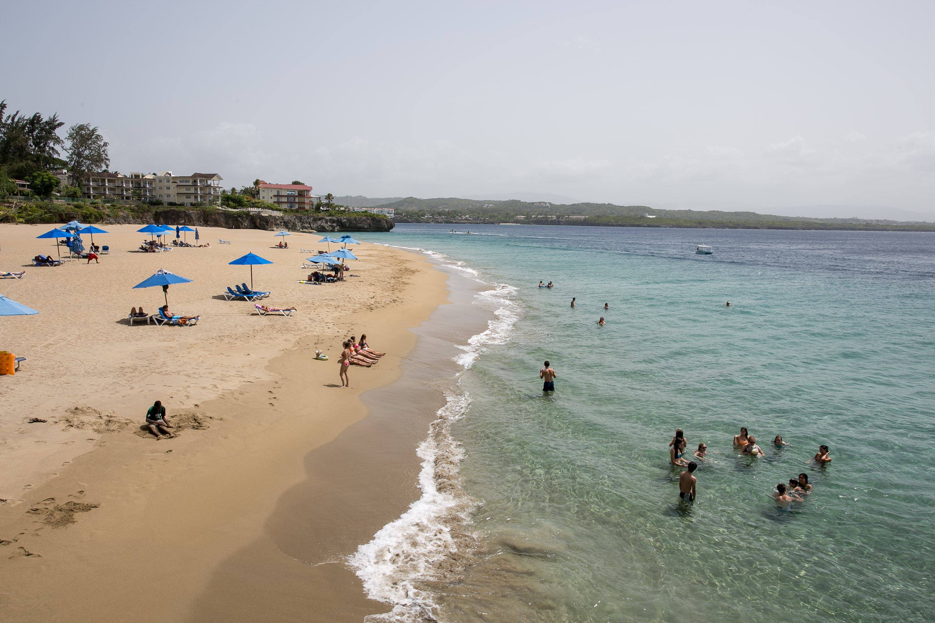 Beach umbrellas line the shore as students wade in the ocean along a curving coastline