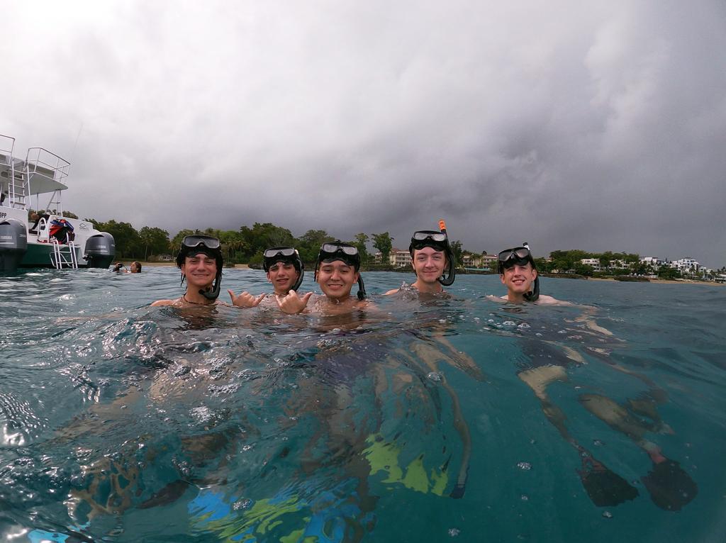 Students wearing snorkel masks pose together in calm coastal waters under a cloudy sky