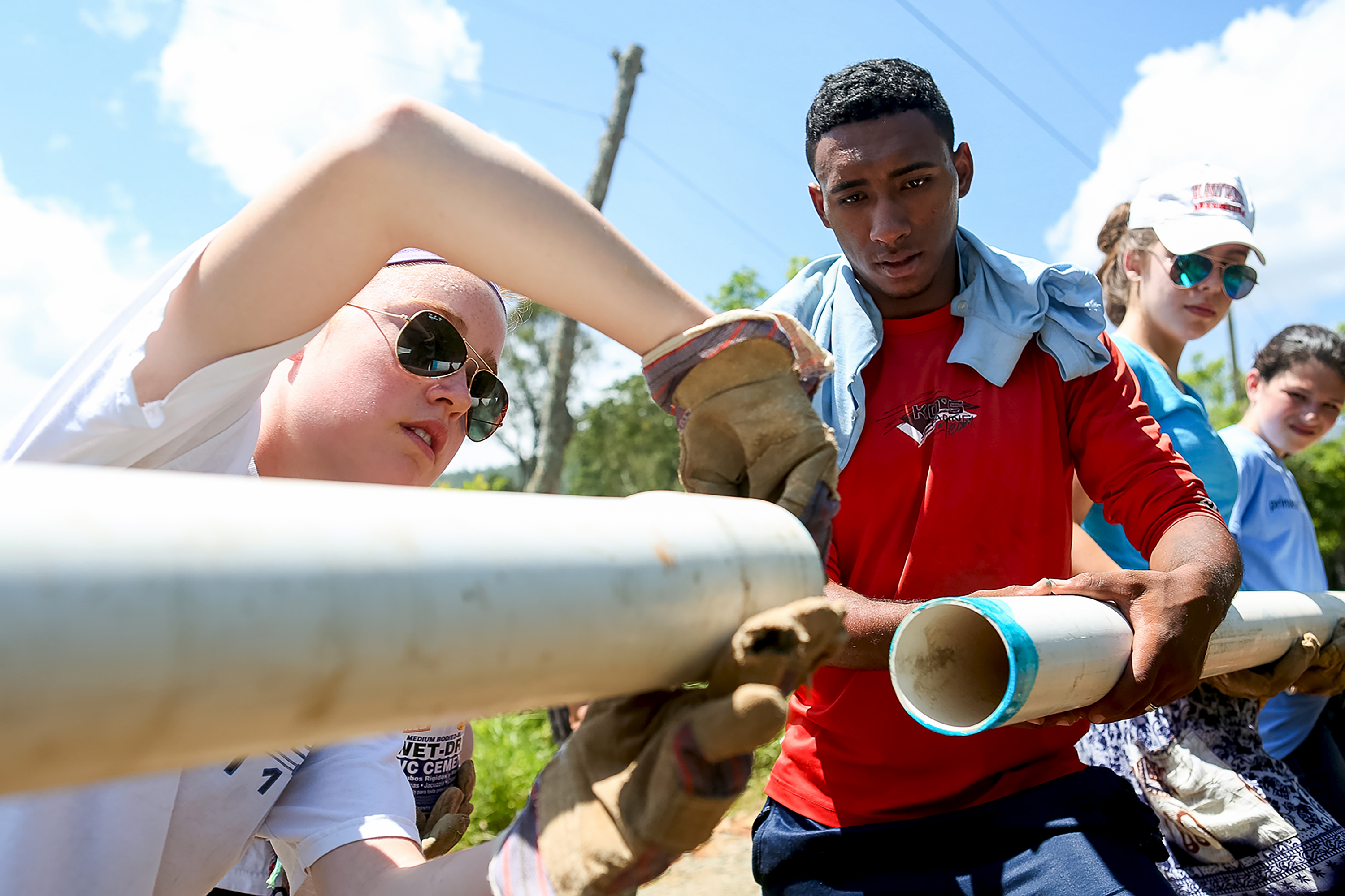 Students wearing work gloves lift PVC pipes at an outdoor service project