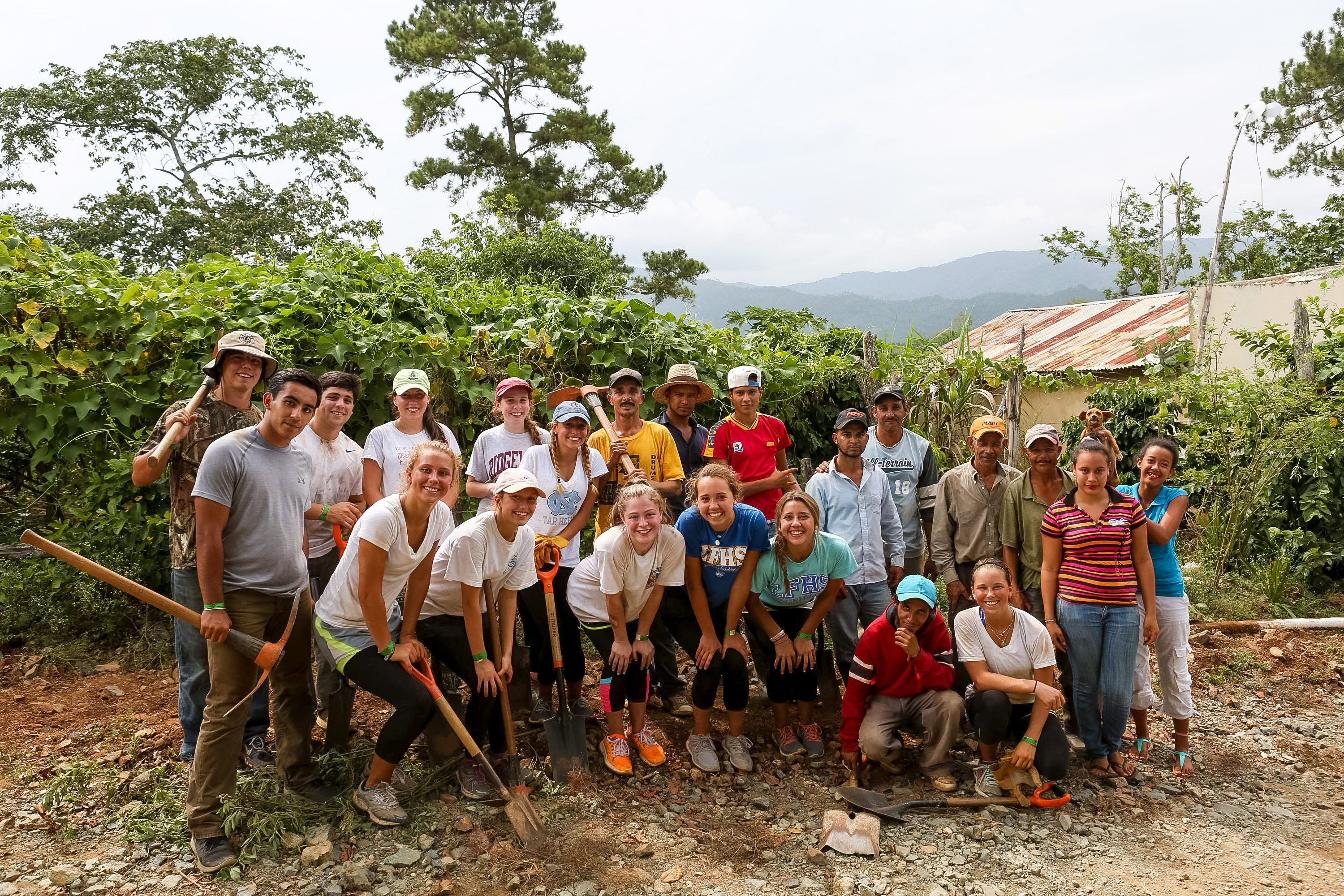 A school trip pauses as students and community members pose with shovels and tools at a mountain village service site