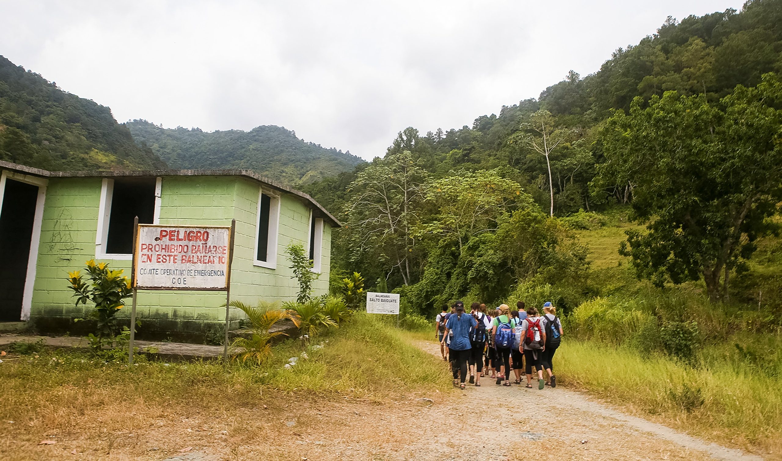 Students hike a dirt road past a green building toward forested mountains