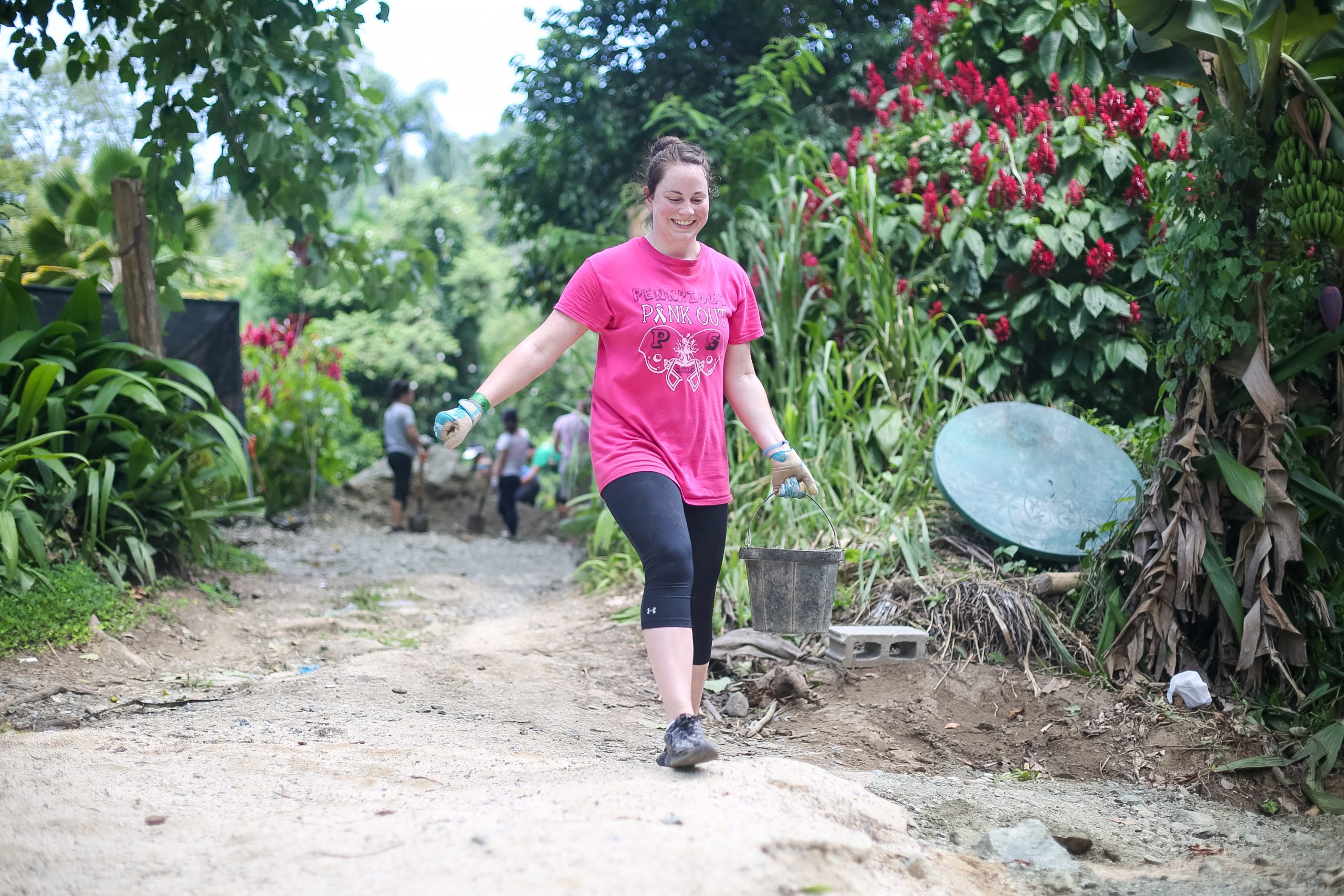 On a school trip, a student walks a dirt path carrying a bucket during a community service project