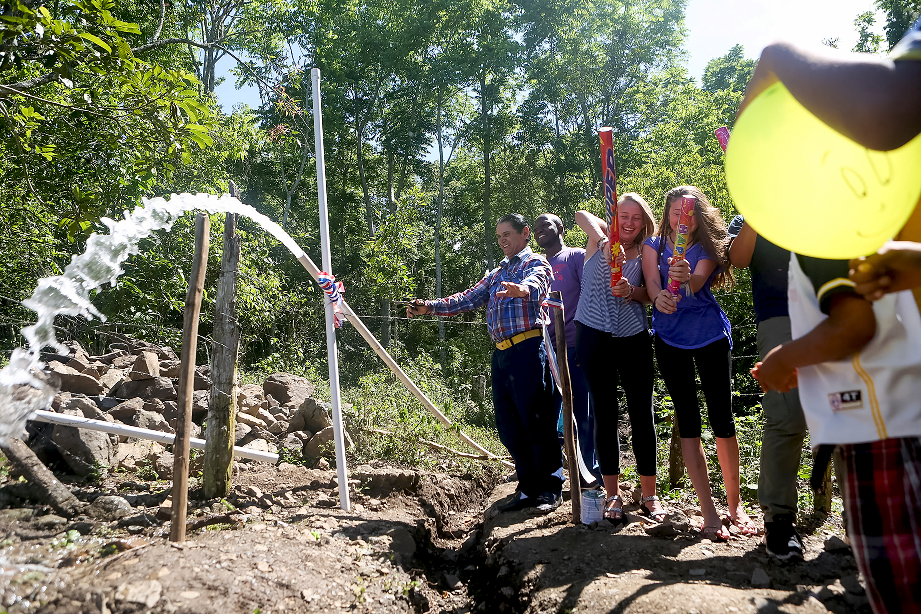 A school trip to Dominican Republic celebrates as water sprays from a newly completed pipeline on a wooded hillside
