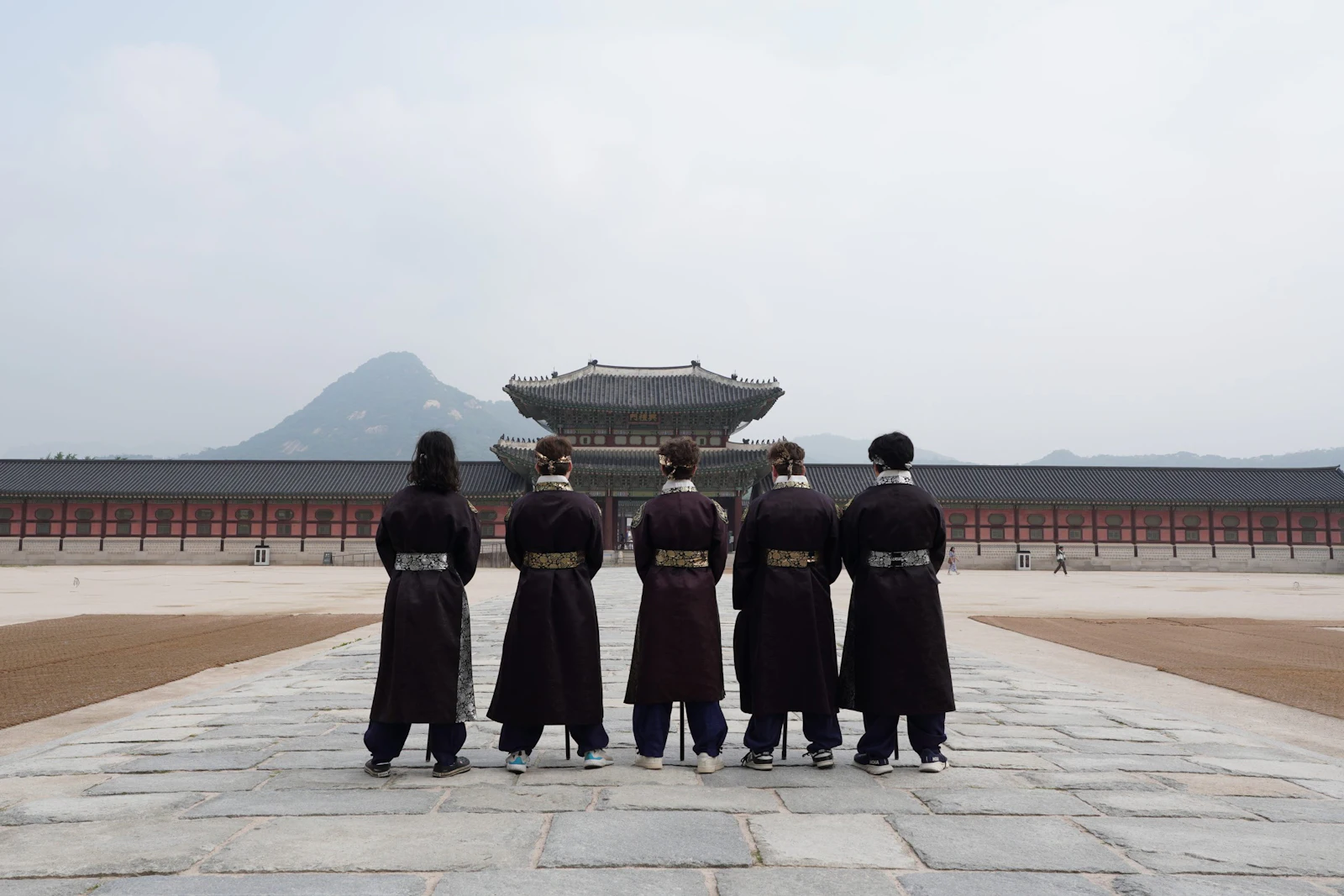 Five people in black traditional robes stand facing a palace hall below a mountain at Gyeongbokgung in Seoul