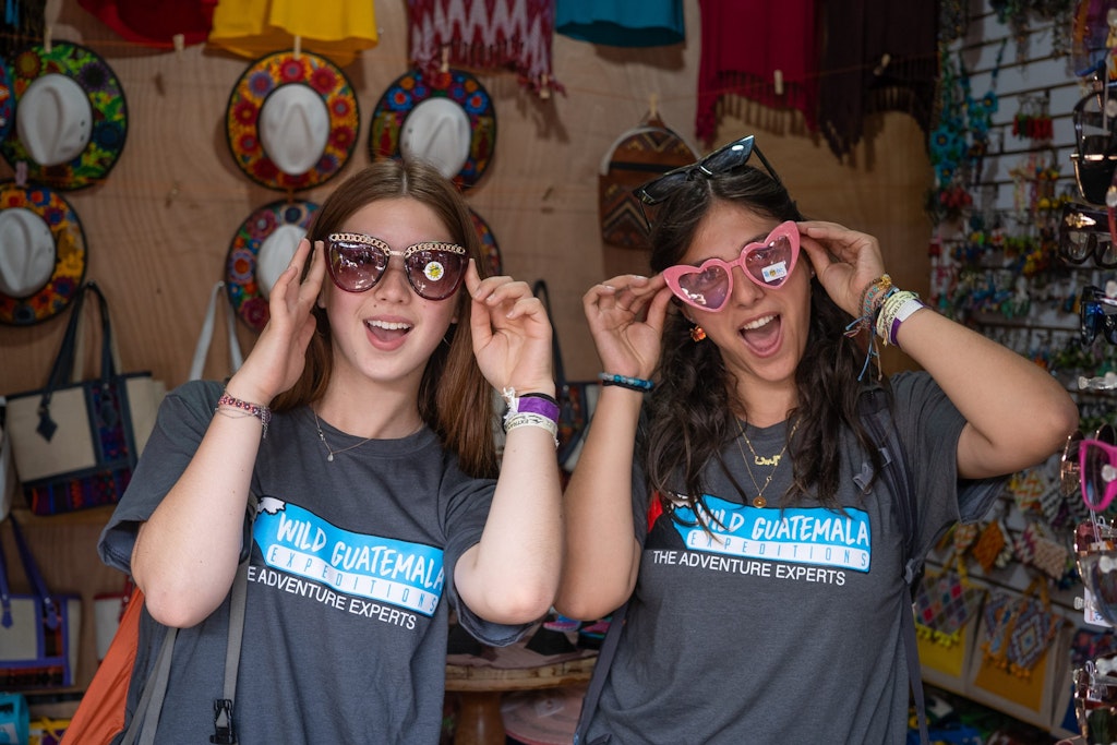 Two student volunteers trying on colorful sunglasses in a local Guatemalan market during a Rustic Pathways volunteer abroad in Central America cultural exchange program.
