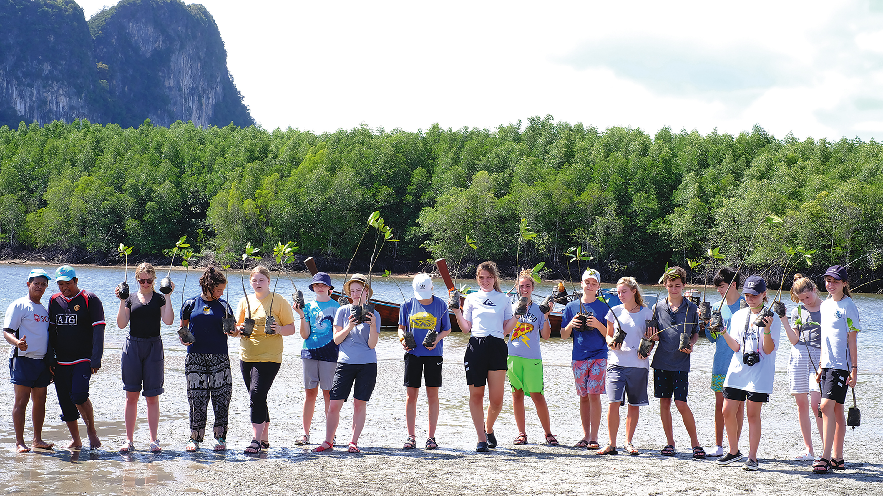 Teens hold mangrove saplings on a muddy shoreline backed by forested limestone cliffs