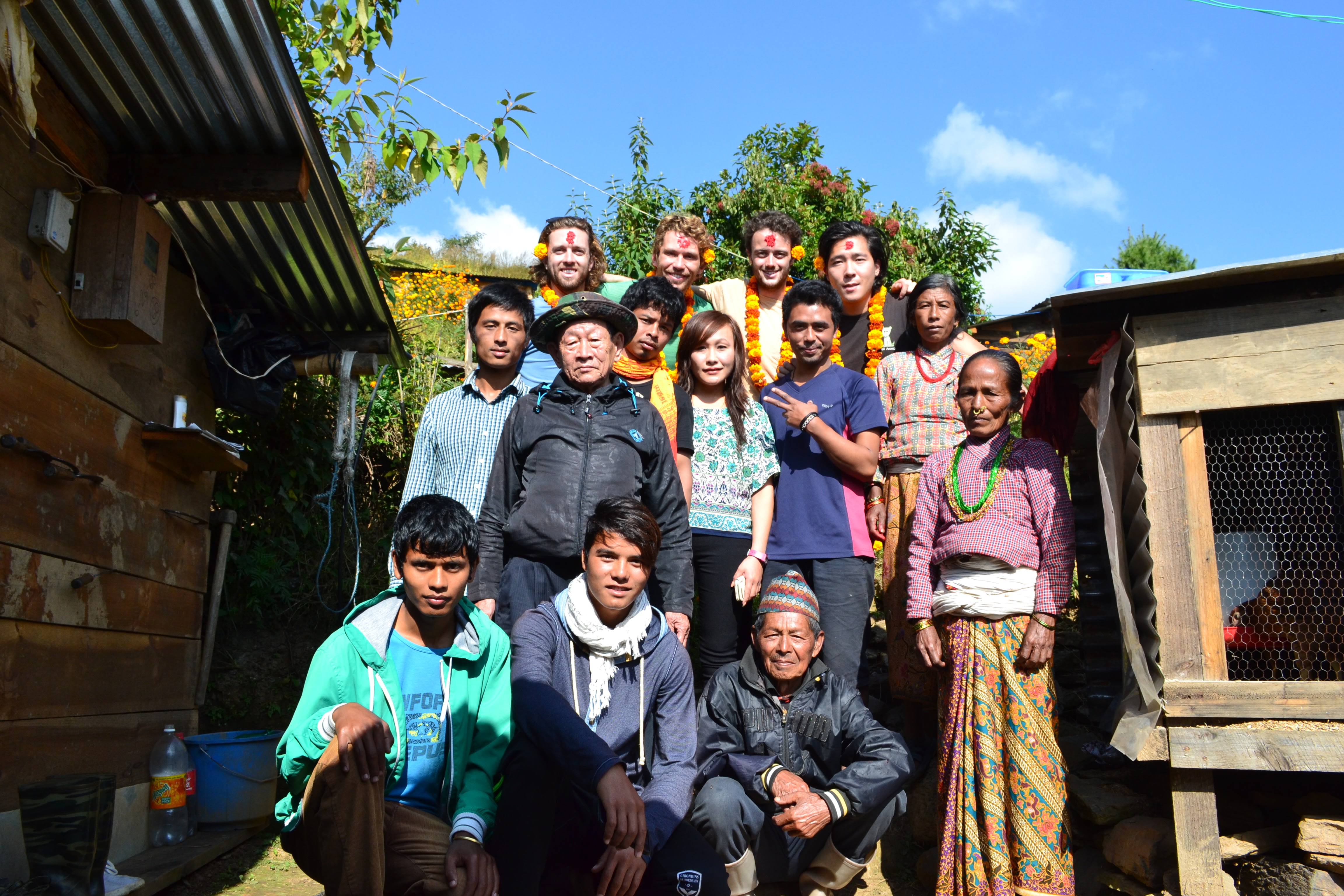 Students and village residents stand together for a group photograph outside a house with a corrugated metal roof.