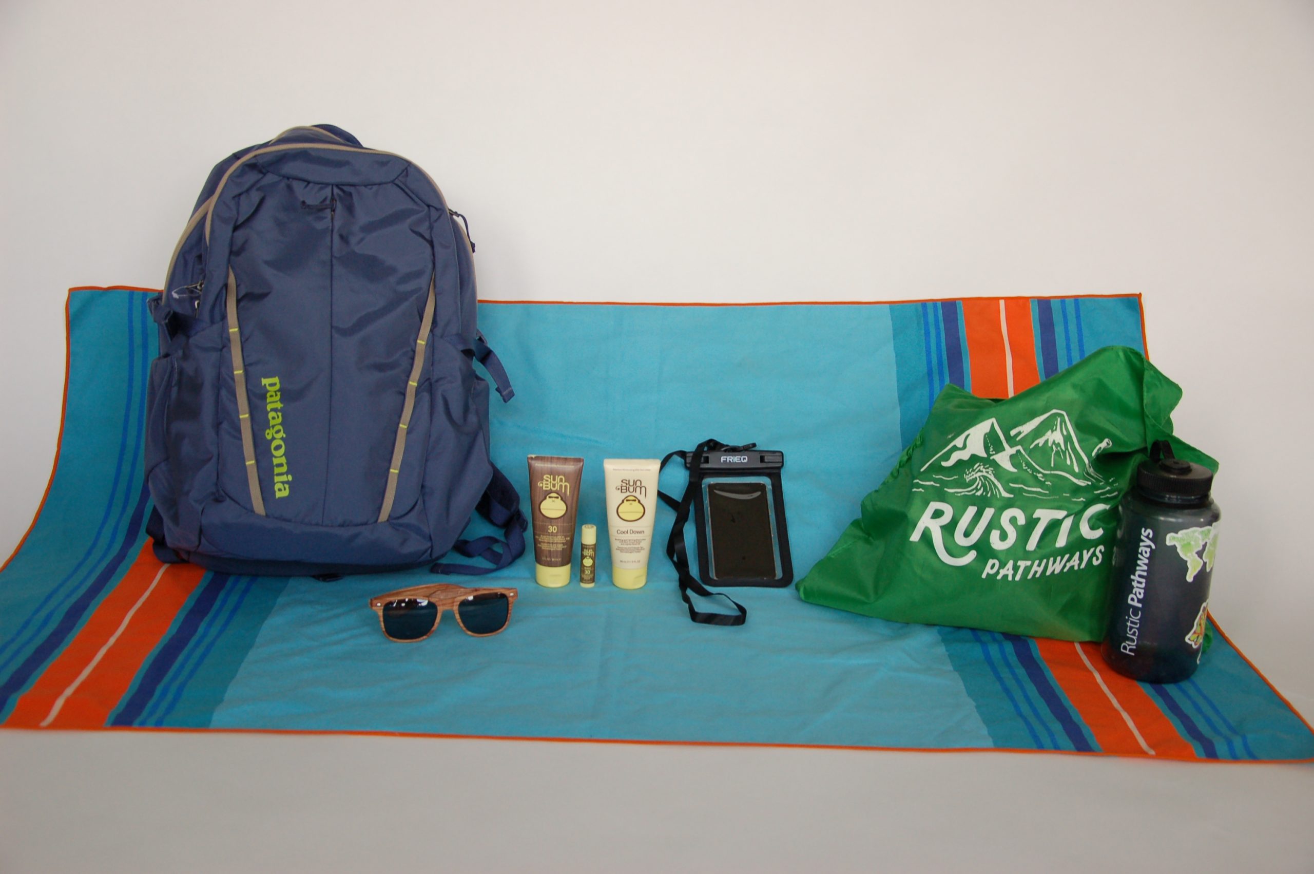 A tarp with a backpack, sunglasses, sunscreen, necklace wallet, and sling back with a Rustic Pathways logo.