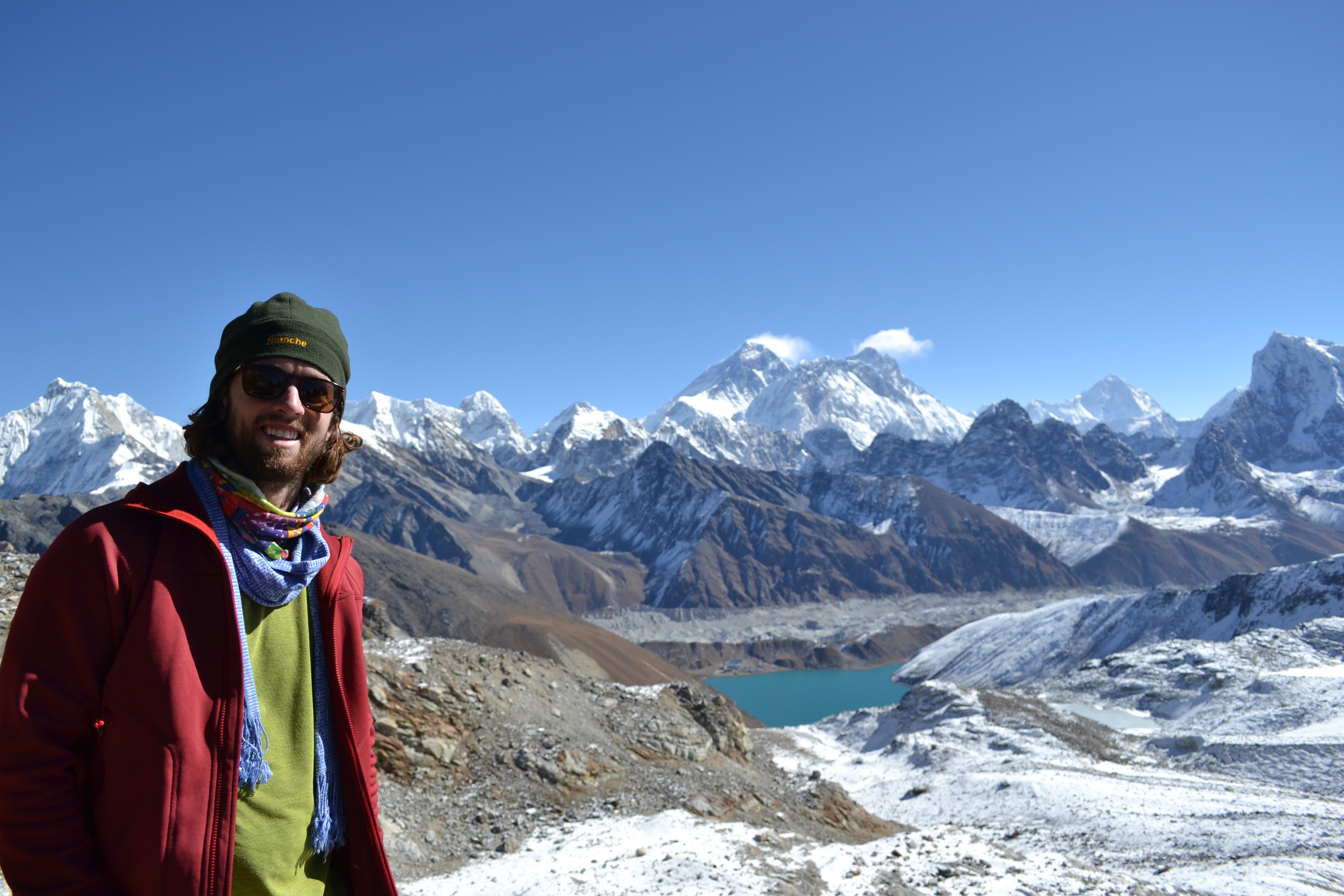 A student poses before a turquoise glacial lake and snow-capped Himalayan peaks